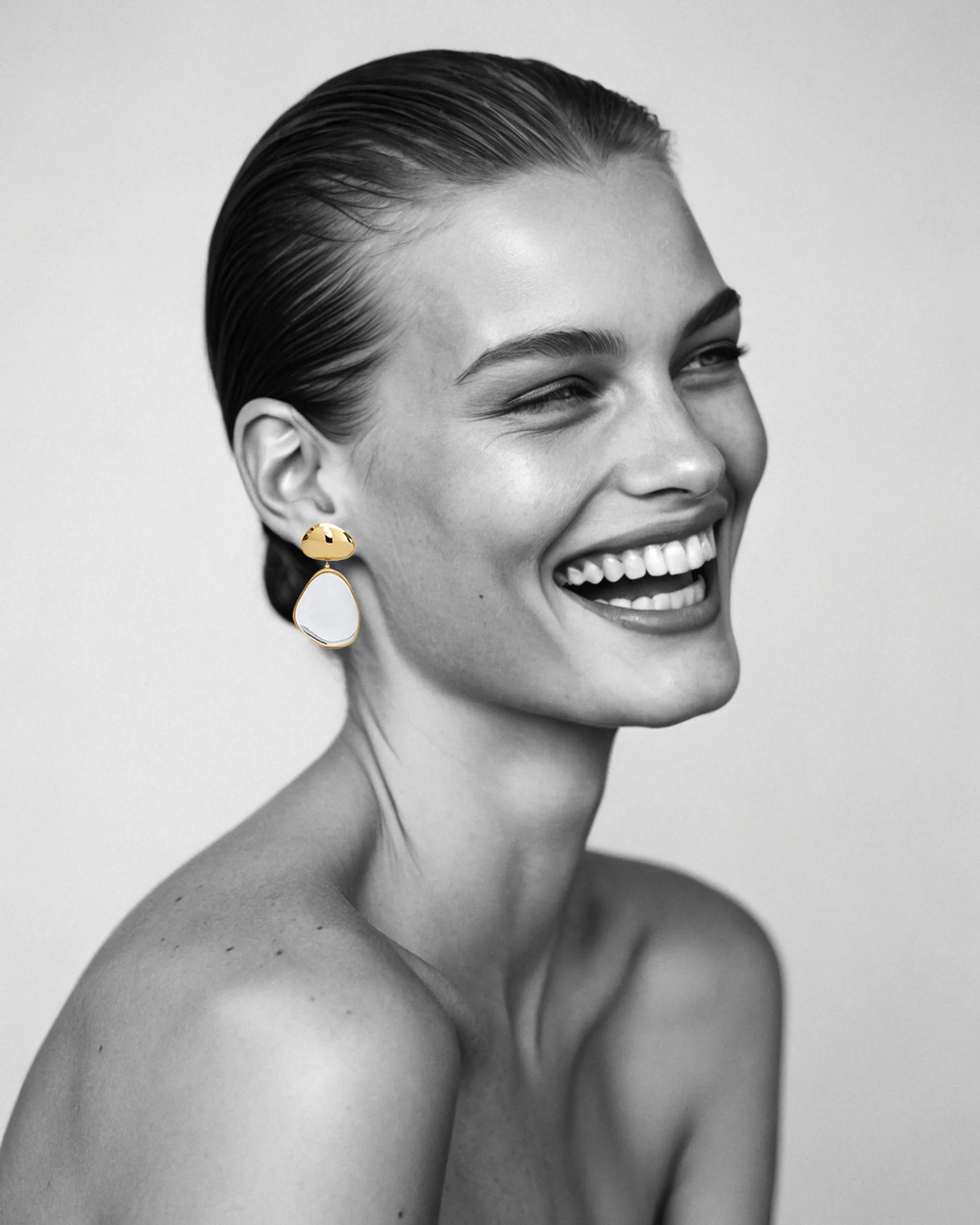 AI image of a smiling model with slicked-back hair wearing a gold and white drop earring, black and white. Alaïa AI Concept.