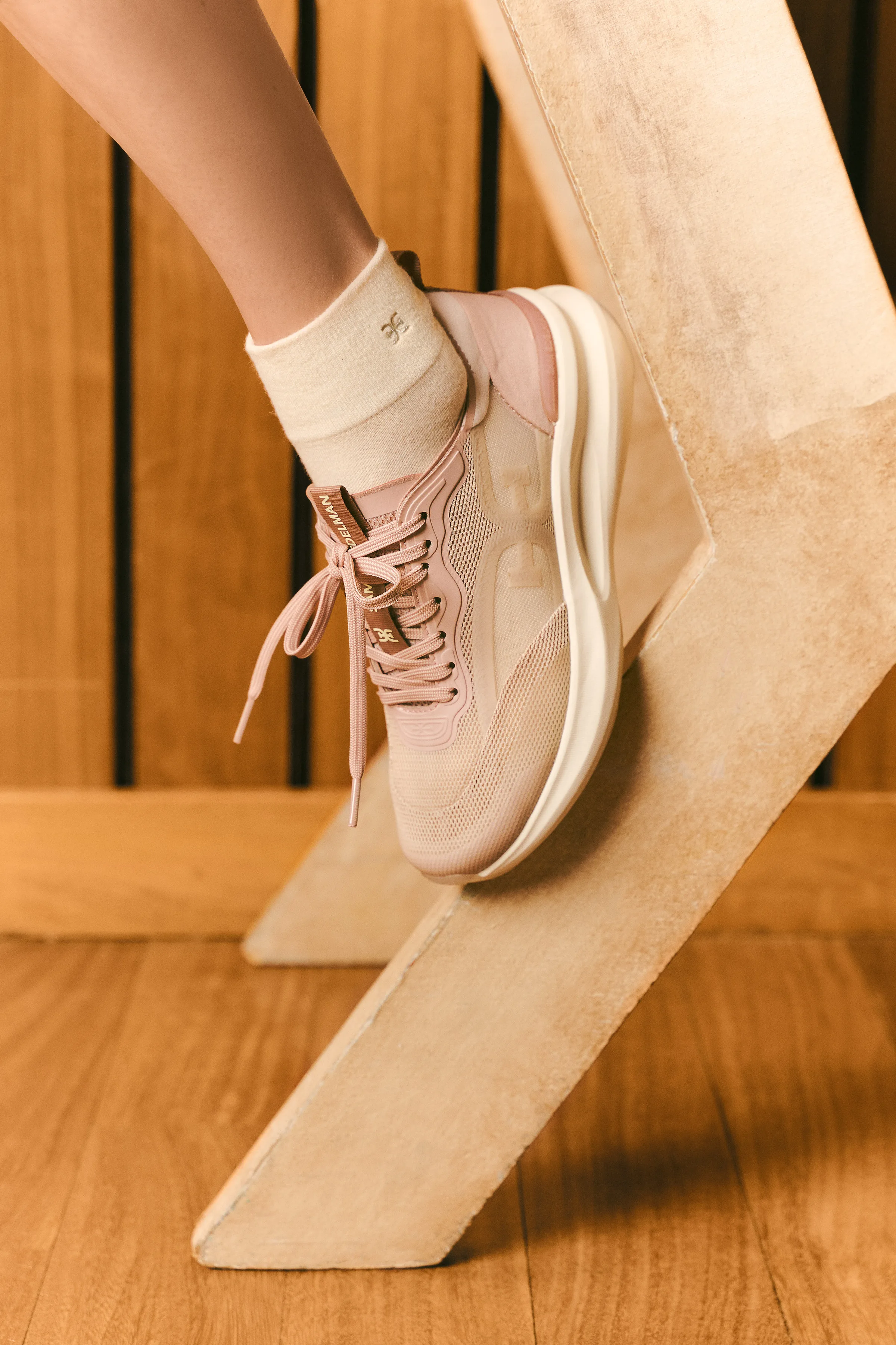 Blush pink mesh sneaker with cream sock resting on a sandstone block against warm wood panels, for Sam Edelman.