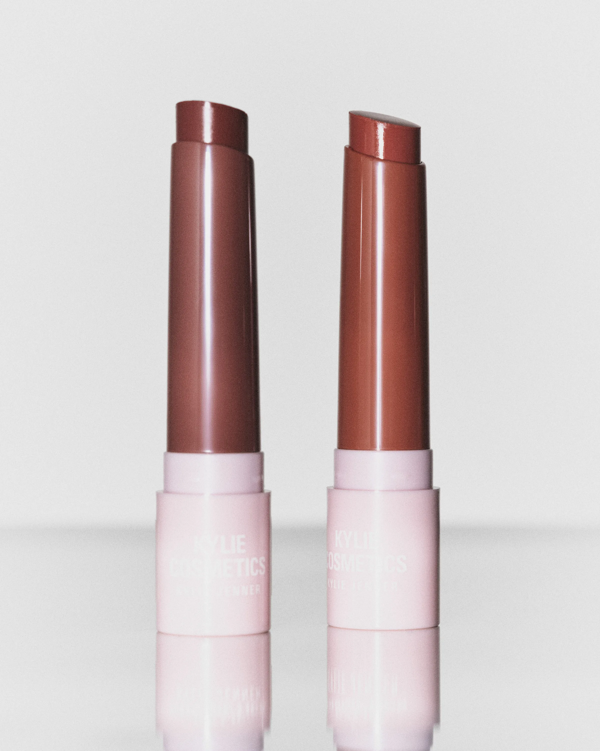 Two uncapped brown lipsticks in pale pink tubes reflected on a glossy surface against a grey backdrop, for Kylie Cosmetics.