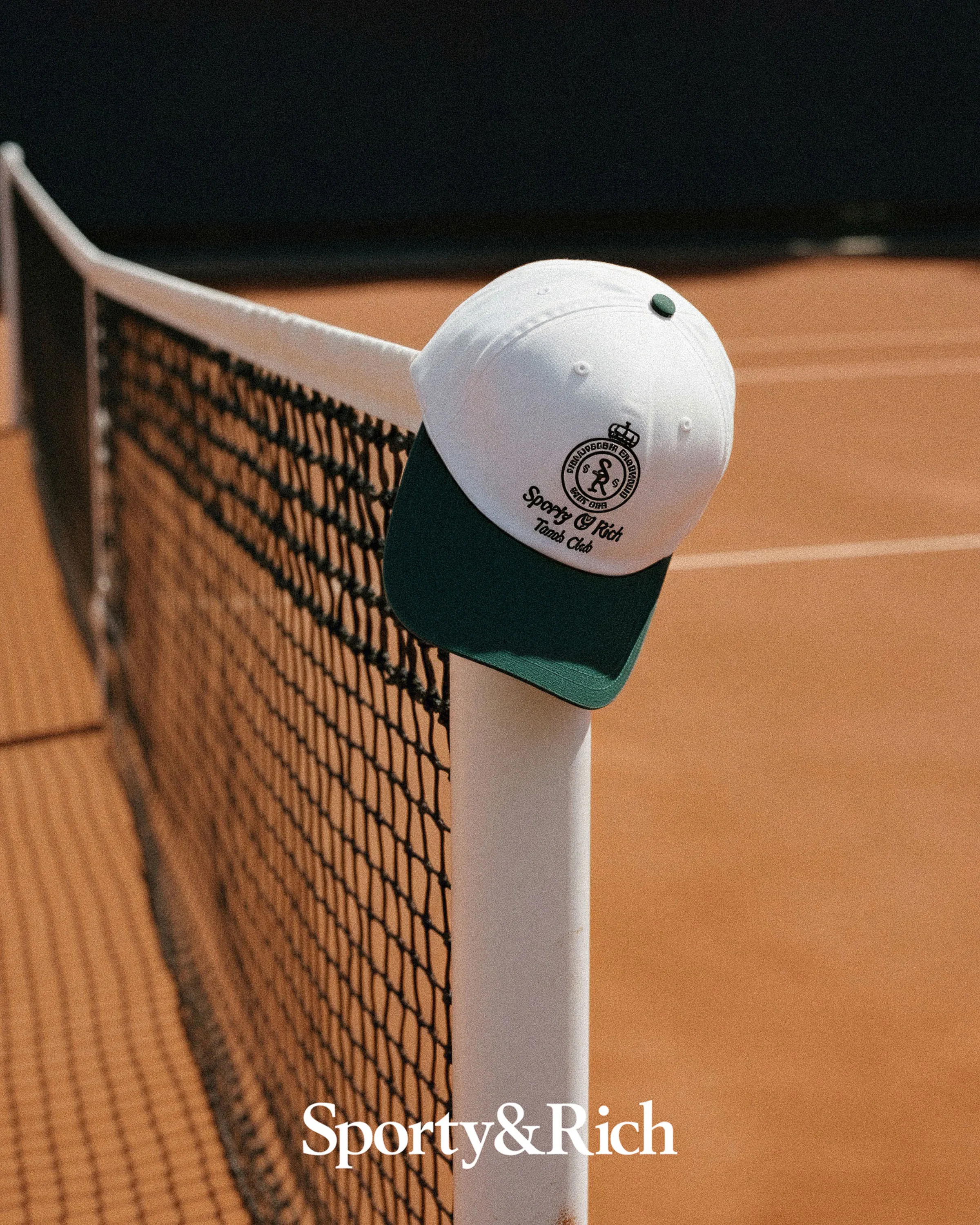 AI image of a white and green tennis club cap hanging on a net post over a clay court. Sporty & Rich AI Concept.