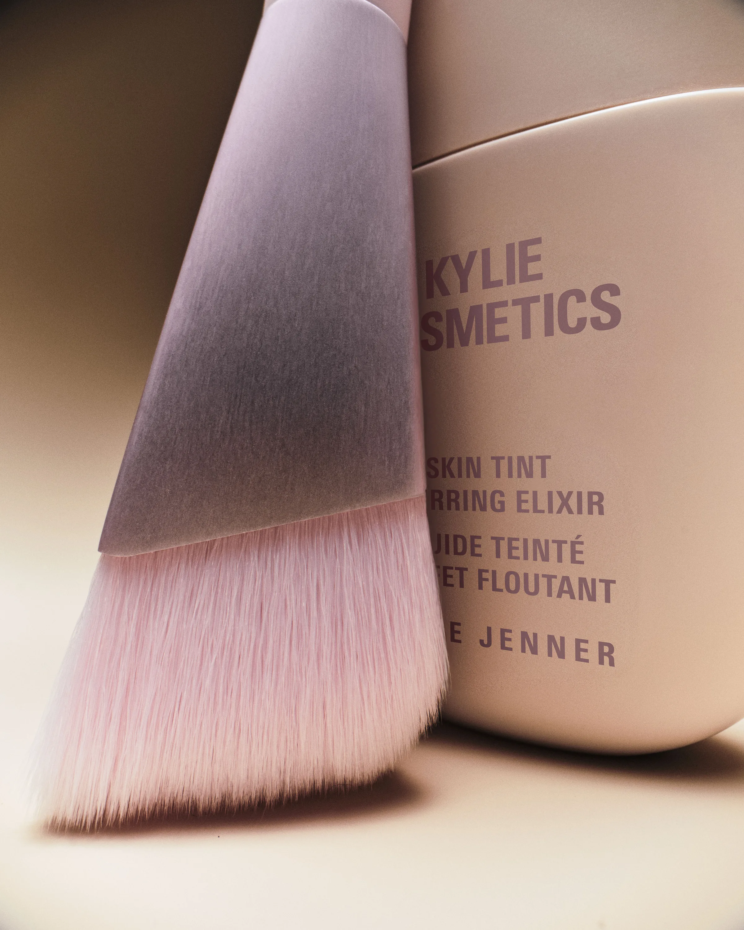 Close-up of a pink-bristled angled foundation brush leaning on a beige Skin Tint Blurring Elixir bottle, for Kylie Cosmetics.
