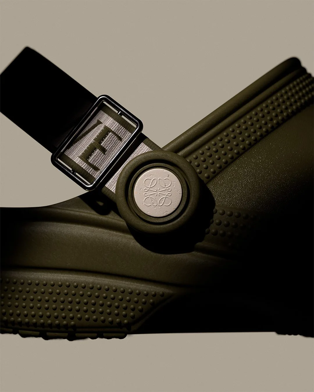 CGI render of an olive green clog with a buckled strap and anagram rivet in close-up on a beige background, for Loewe.