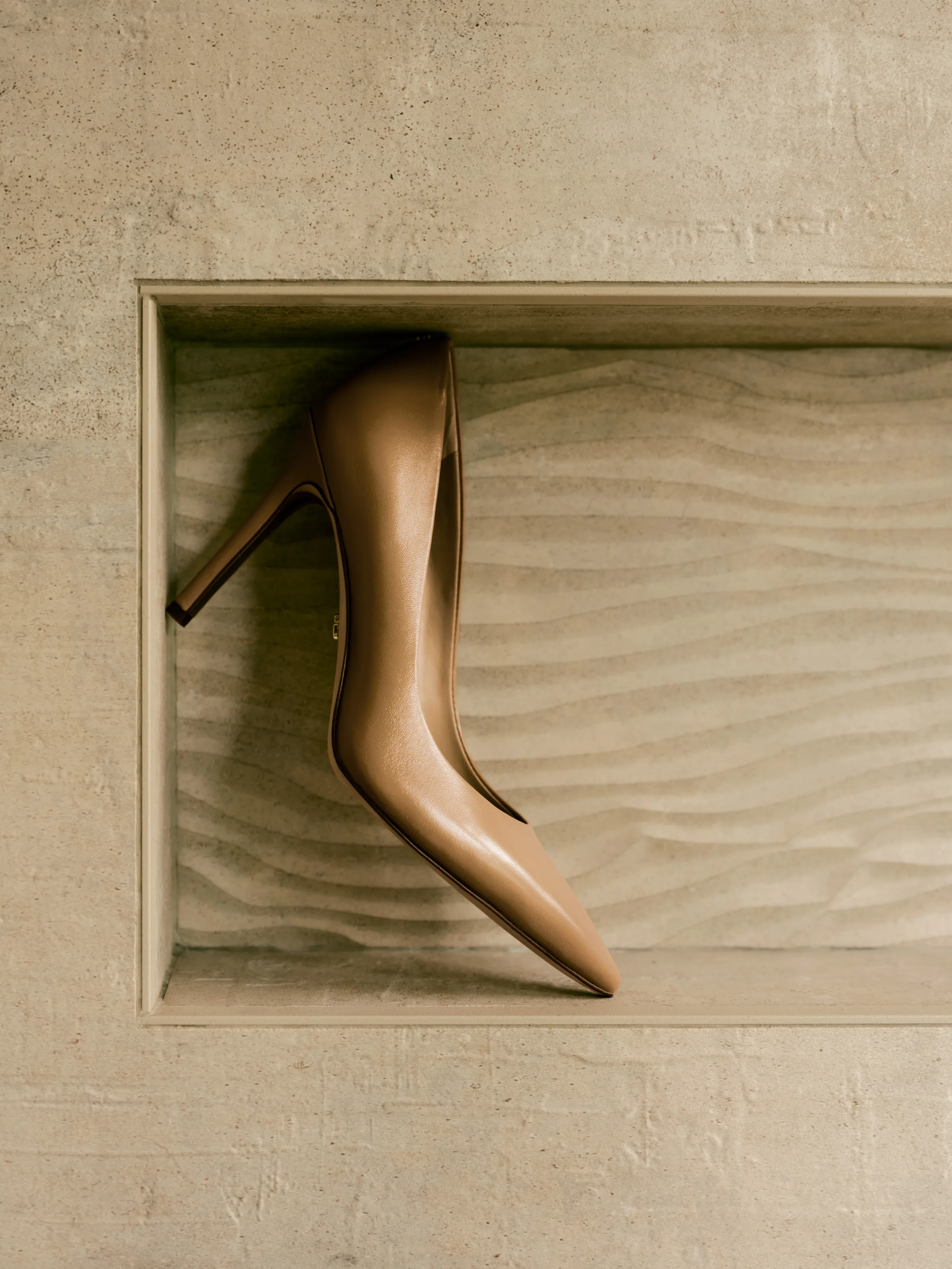 Tan leather pointed-toe pump balanced in a sandstone wall niche with rippled sand texture, for Sam Edelman.