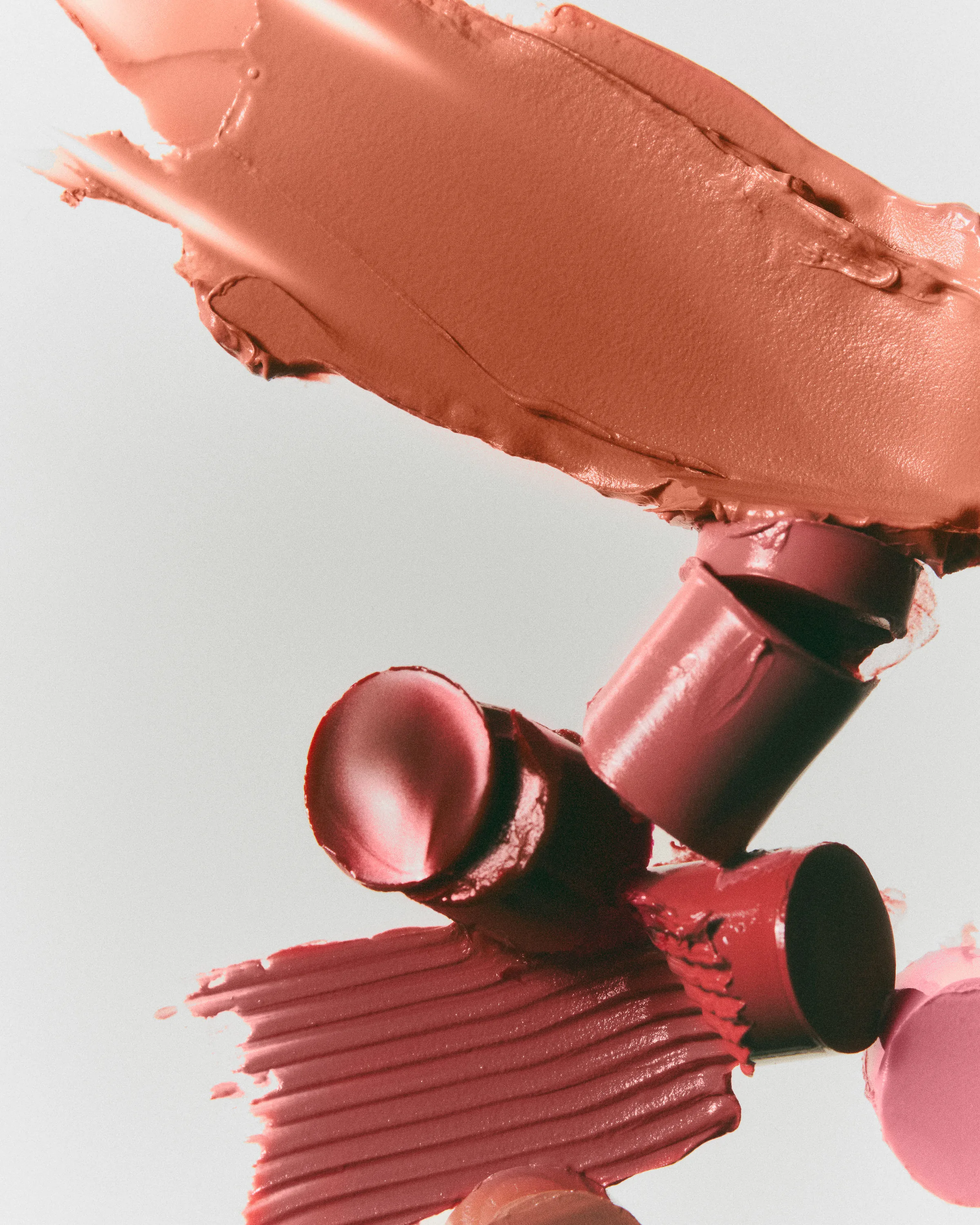 Still life of cut lipstick bullets and smeared swatches in coral, rose and pink on a pale grey background, for Kylie Cosmetics.