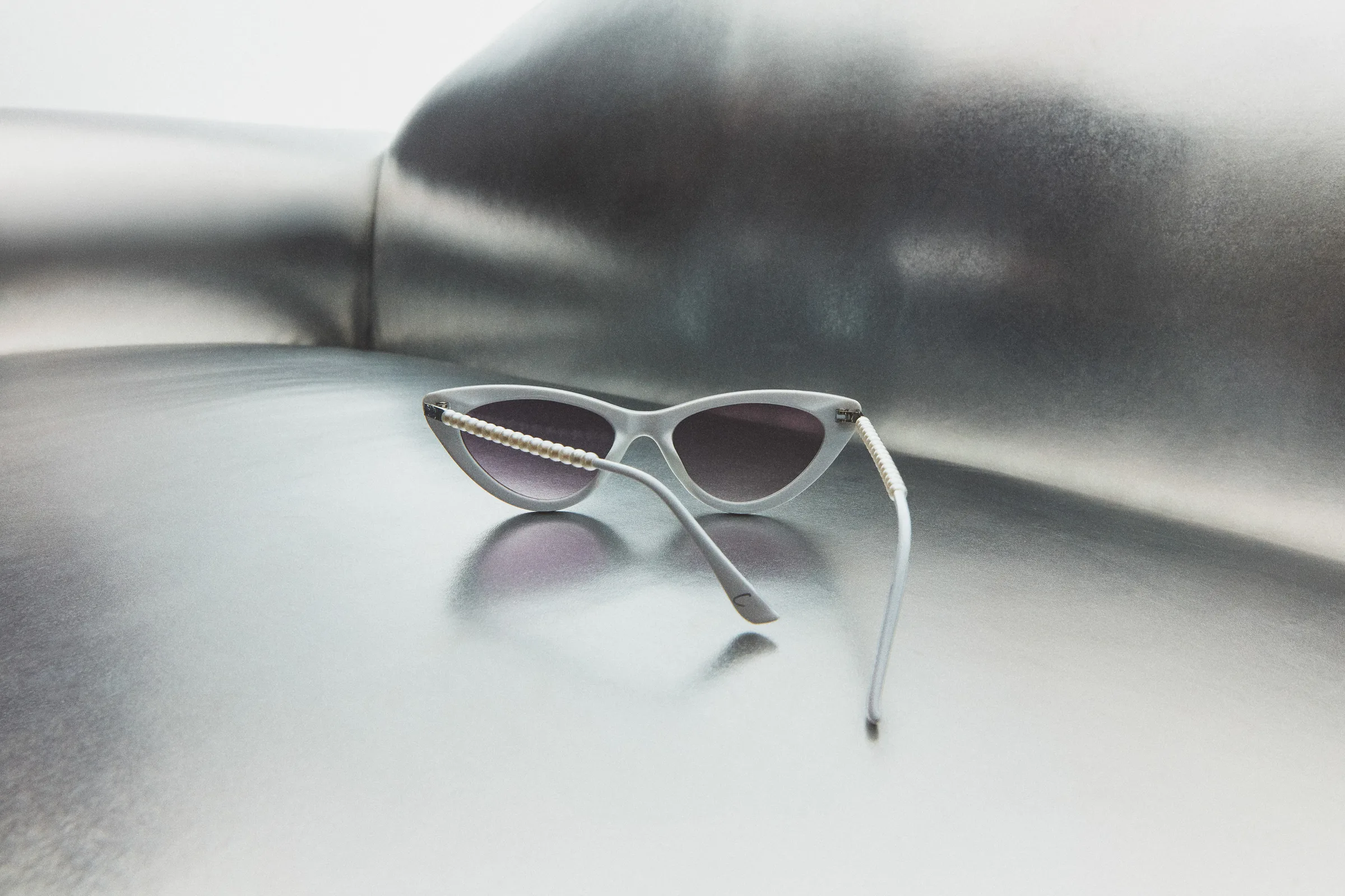 White cat-eye sunglasses with pearl-trimmed arms and gradient lenses on a brushed silver metallic surface, for Sam Edelman.