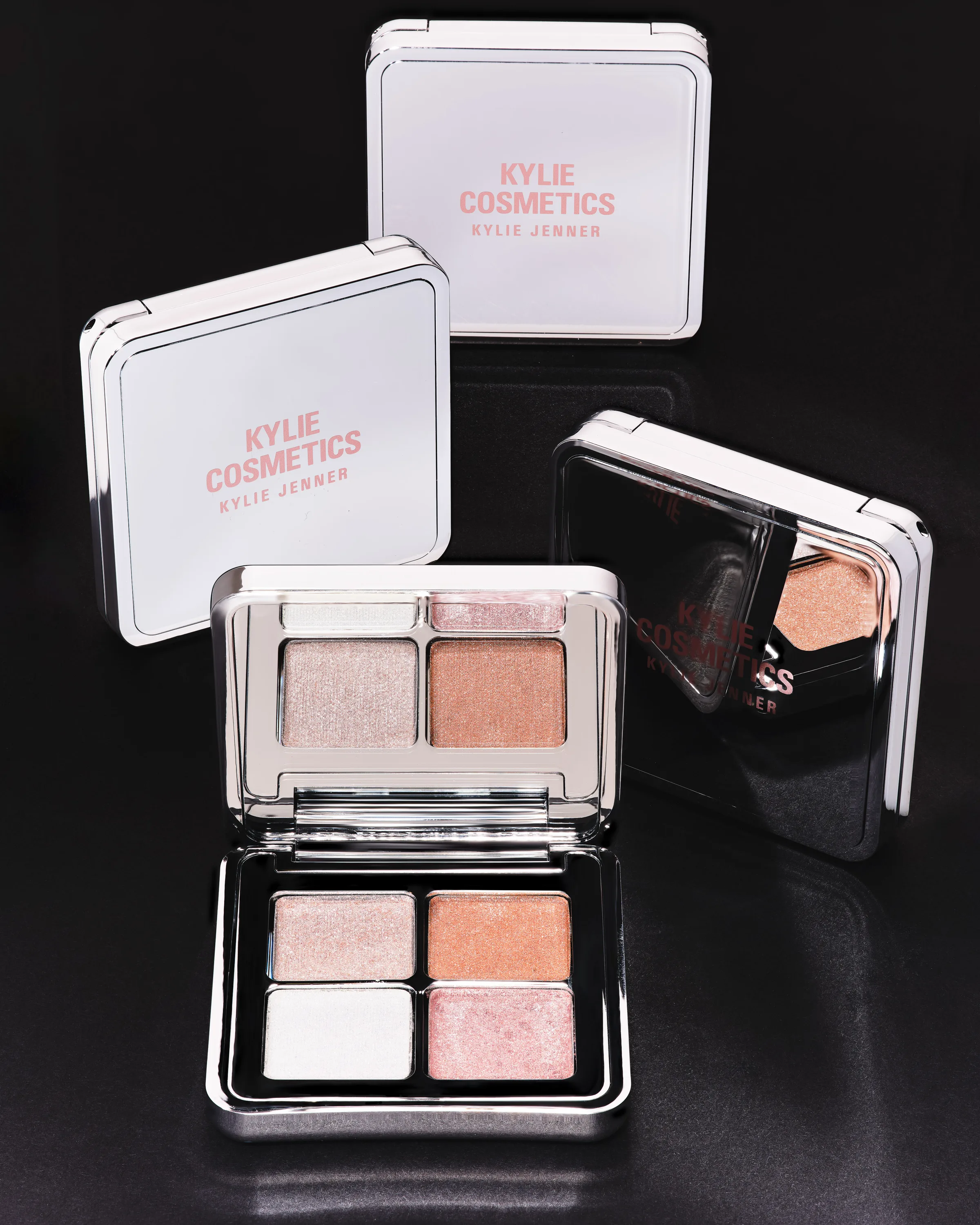Silver mirrored eyeshadow quad compacts, one open showing shimmering champagne and peach shades, on black, for Kylie Cosmetics.