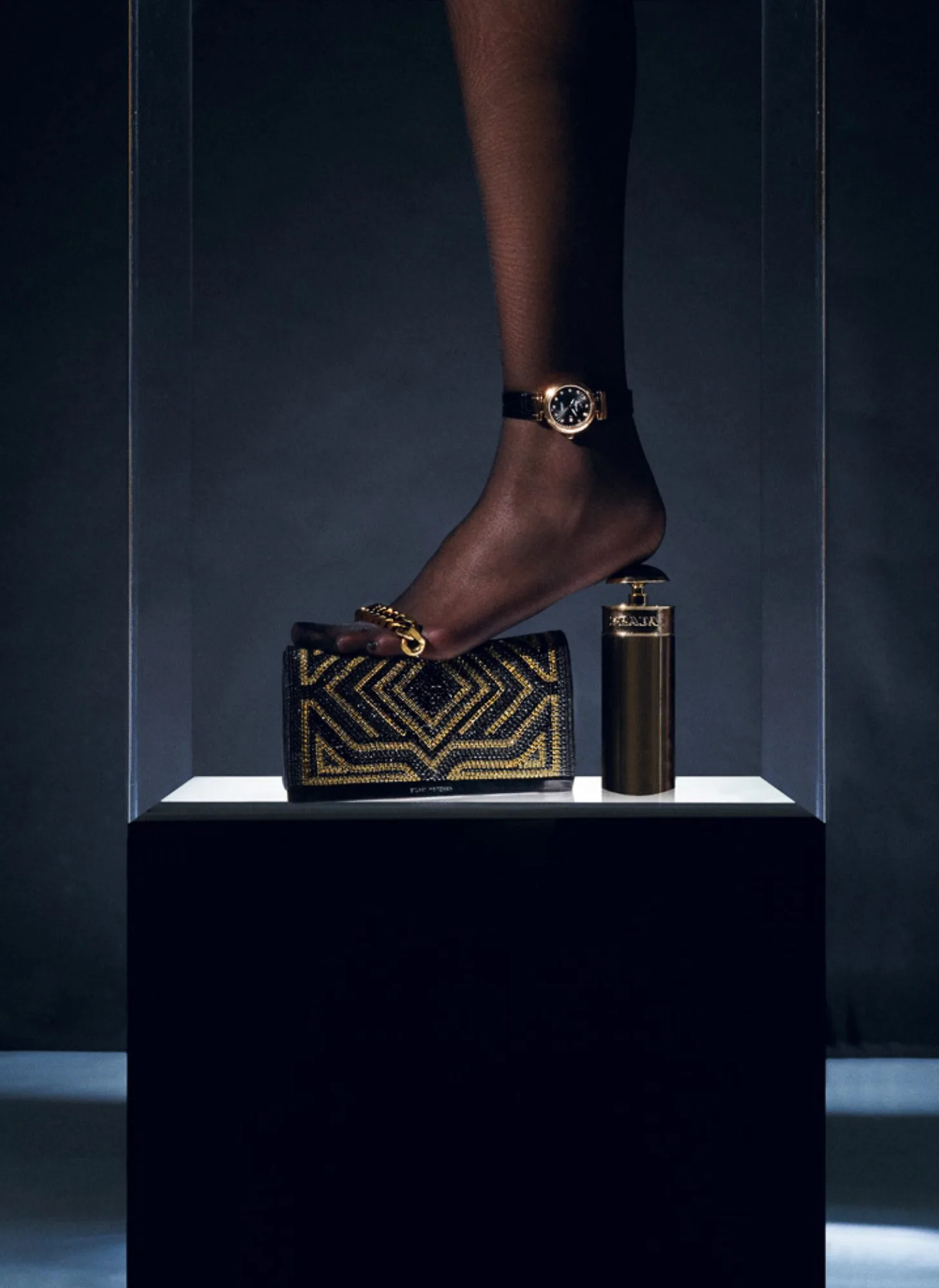 Stockinged foot with gold watch anklet resting on a gold-studded clutch and perfume bottle atop a lit plinth, for Interview Magazine.