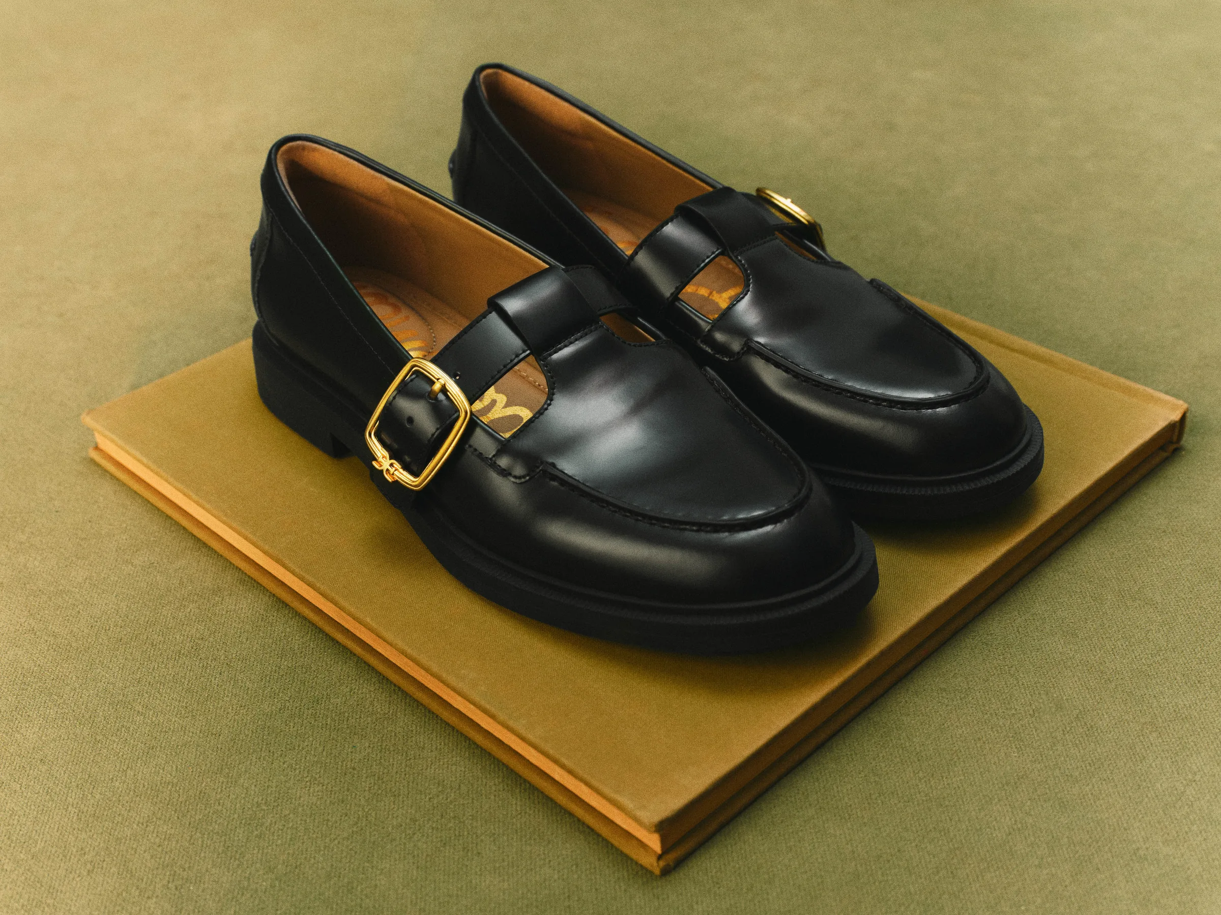 Black leather T-strap loafers with gold buckles resting on an ochre book against an olive backdrop, for Sam Edelman.
