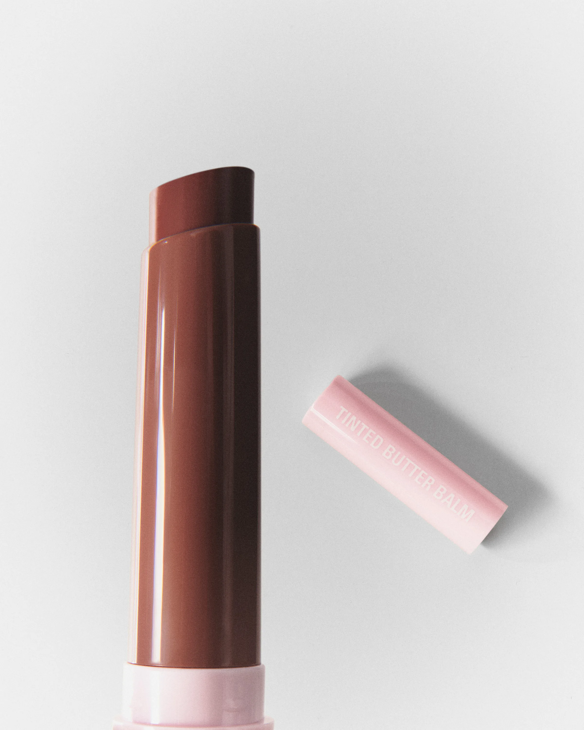 Mocha Kiss tinted butter balm uncapped beside its pink cap on a cream background, still life, for Kylie Cosmetics.