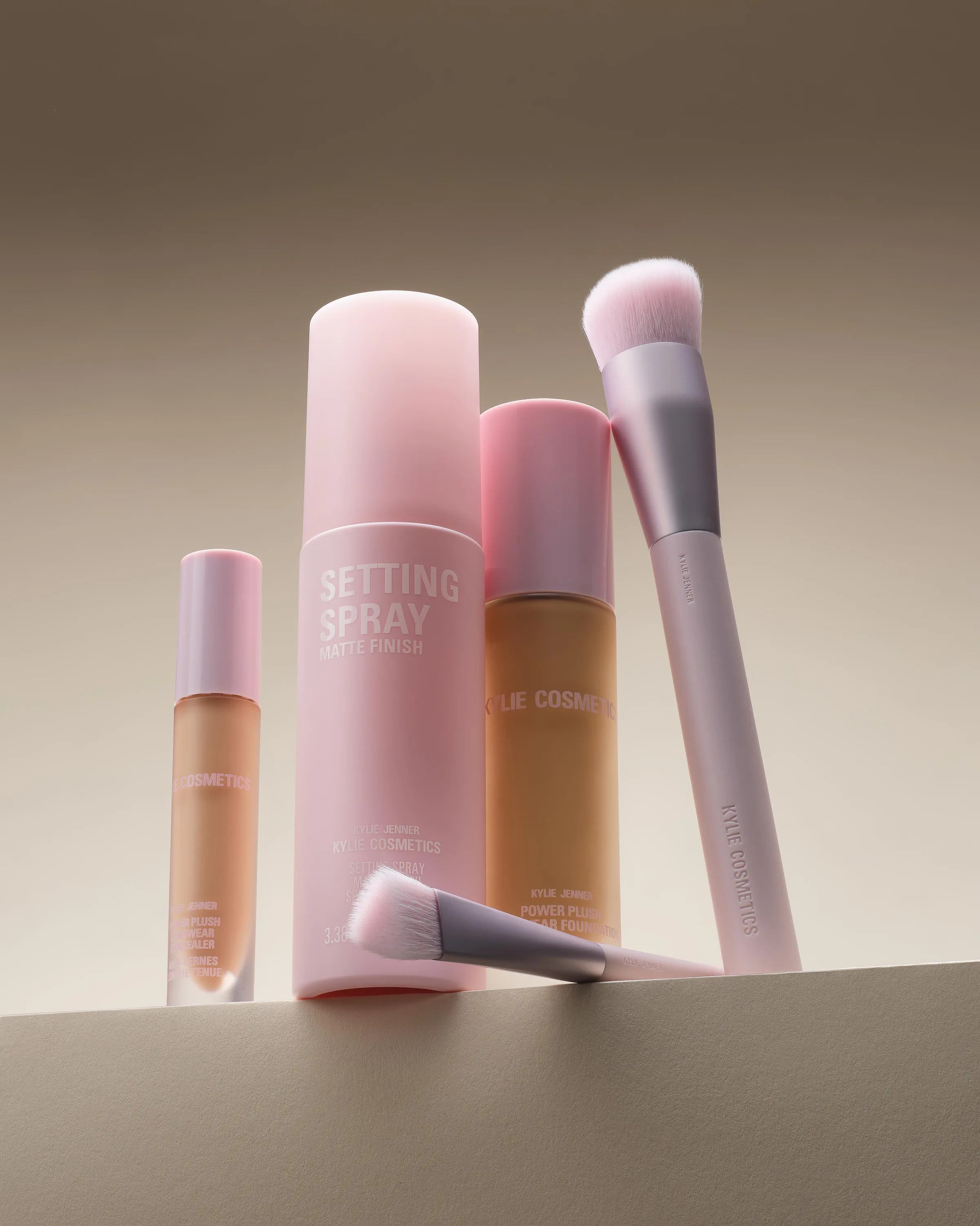 Pink setting spray, foundation, concealer and makeup brushes arranged on a beige ledge, for Kylie Cosmetics.