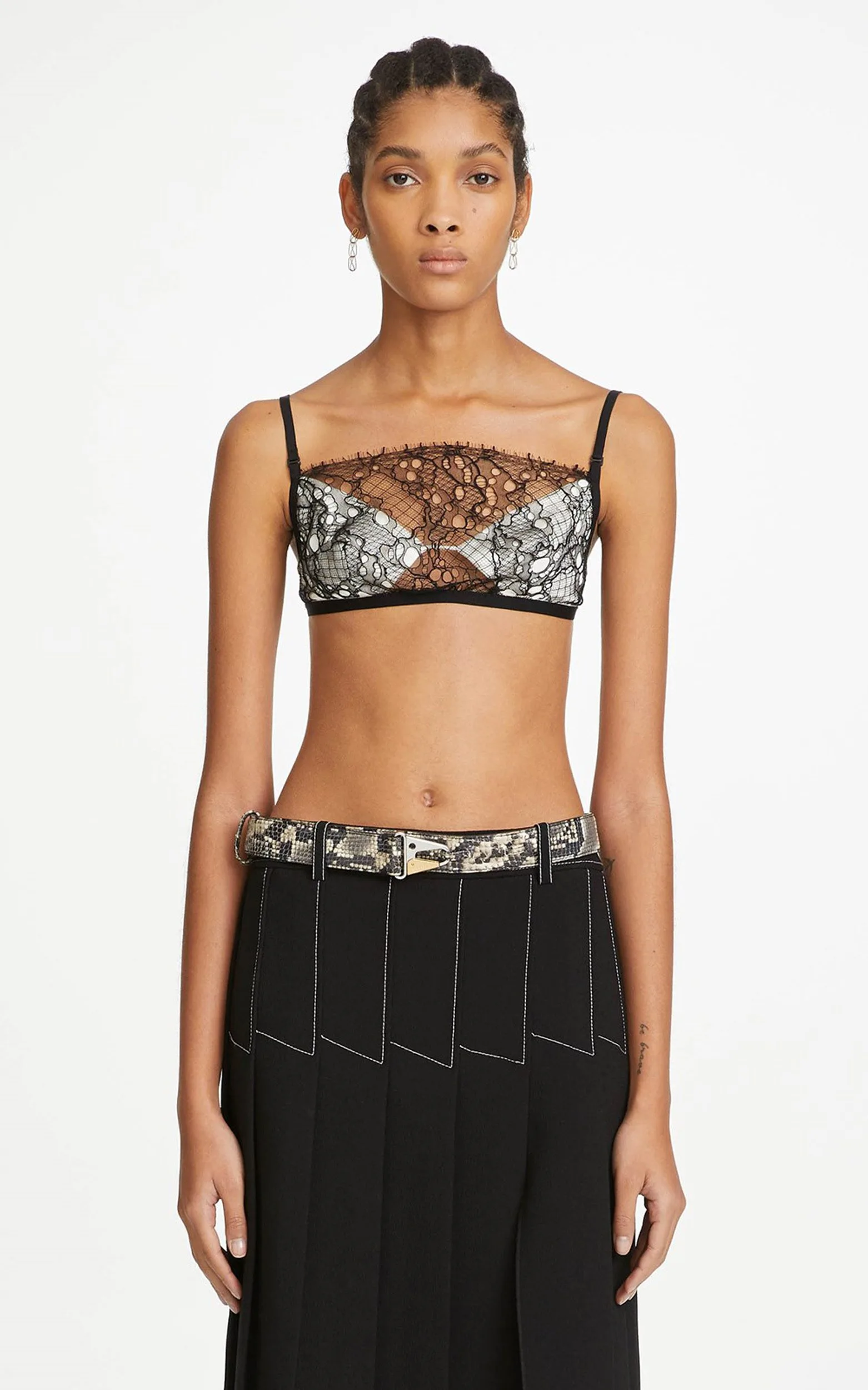 Model in a black and white lace bralette, snakeskin belt and black pleated skirt on a white background, for Dion Lee.
