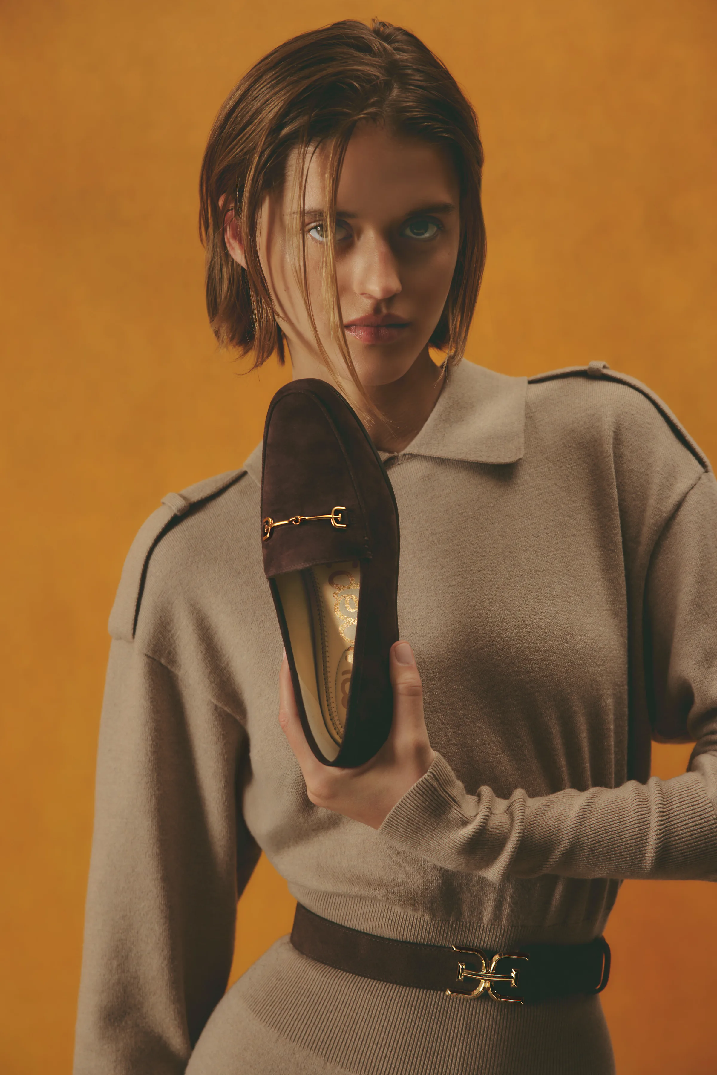 Model in a taupe knit dress holds a brown suede loafer with gold bit hardware against an amber backdrop, for Sam Edelman.