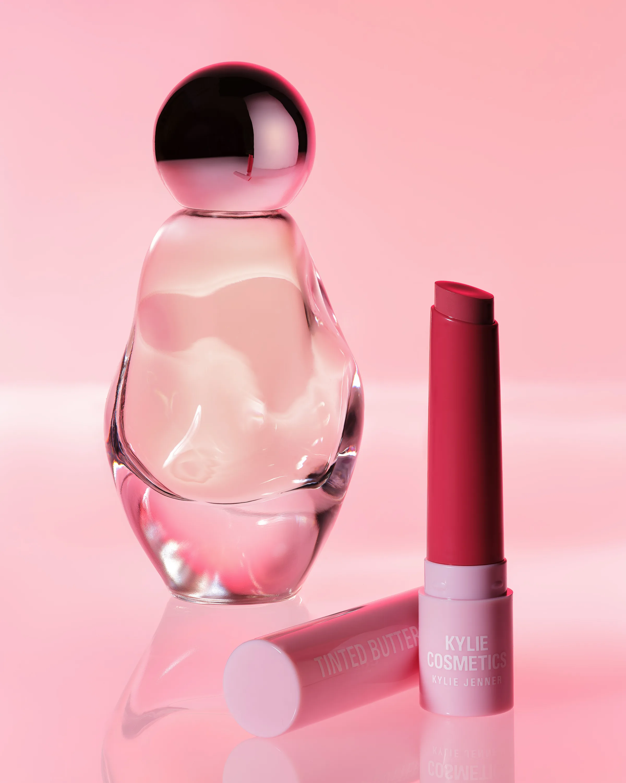 Clear sculpted Cosmic perfume bottle beside a raspberry Tinted Butter Balm on a glossy pink backdrop, for Kylie Cosmetics.
