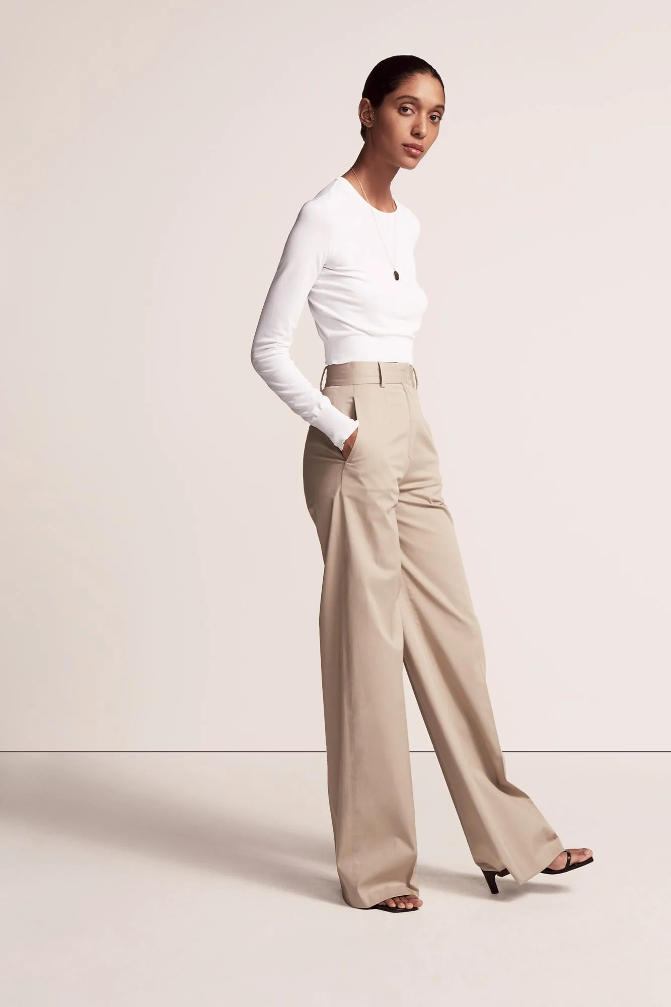 Model in a white long-sleeve top and khaki wide-leg trousers with black heeled sandals on a cream set, for Another Tomorrow.