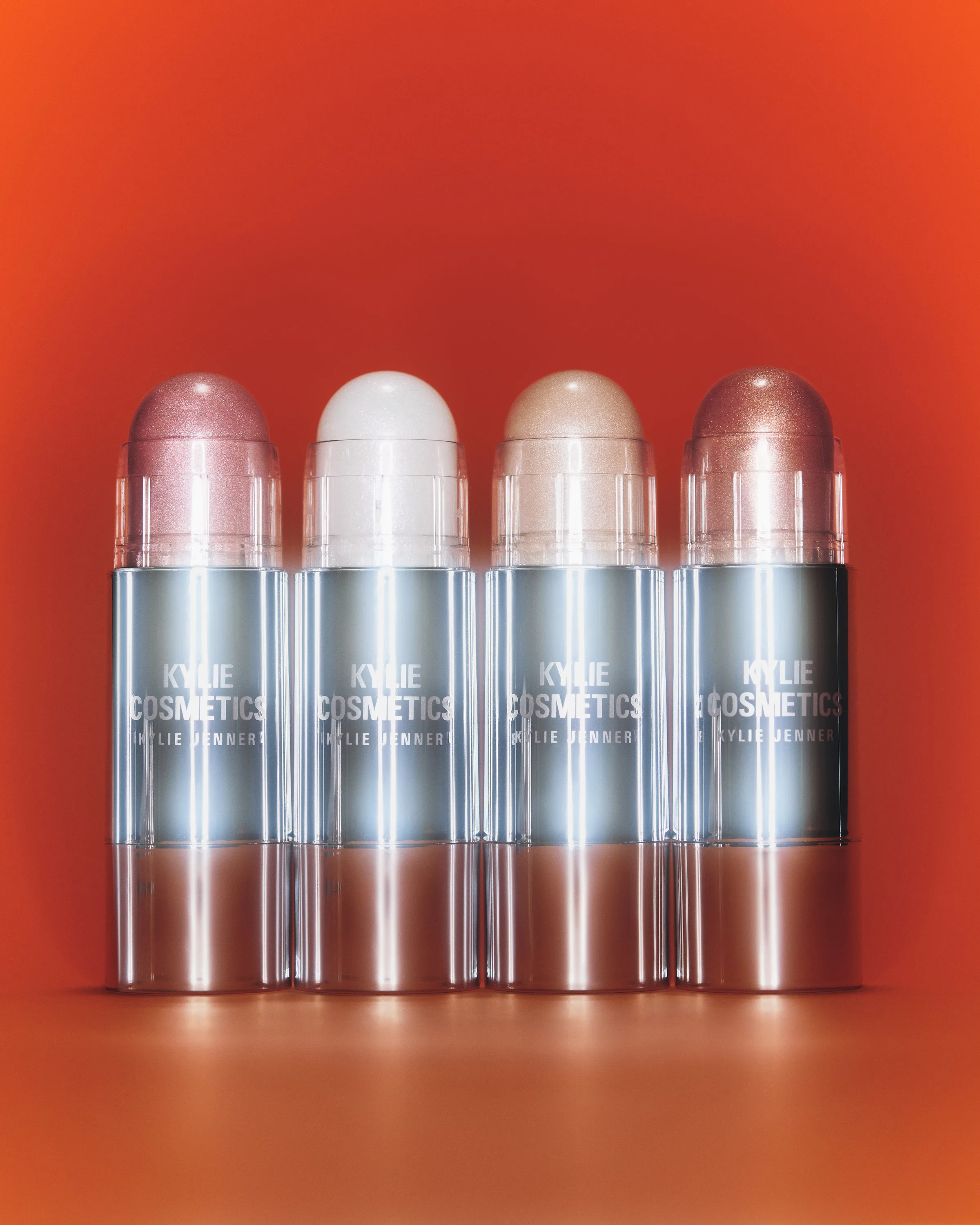 Four shimmering dewy balm sticks in silver tubes lined up against a glowing orange background, for Kylie Cosmetics.