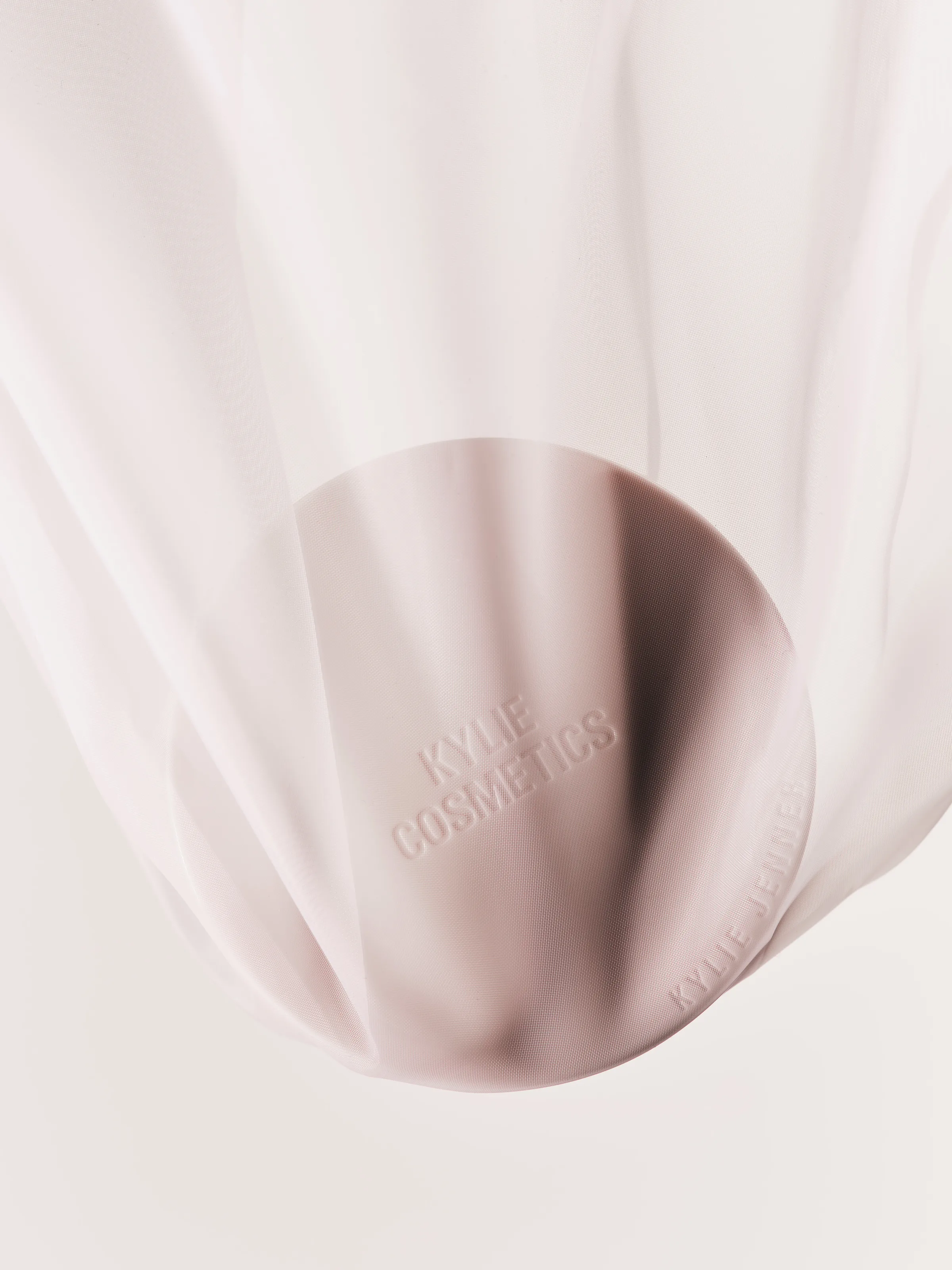 Pale pink embossed compact draped in sheer white mesh fabric on a blush background, for Kylie Cosmetics.