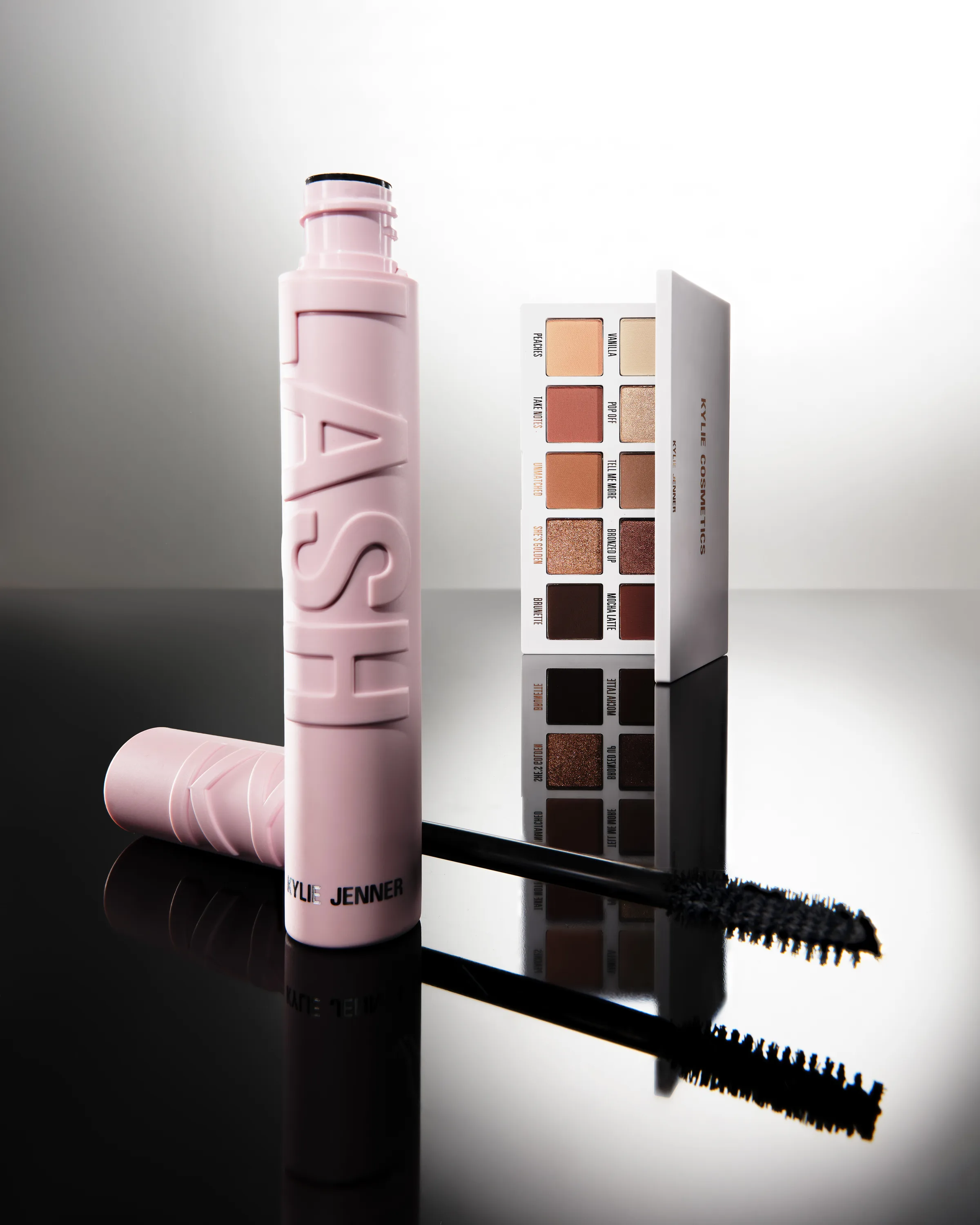 Pink Kylash mascara tube and wand beside an open bronze eyeshadow palette on a reflective black surface, for Kylie Cosmetics.