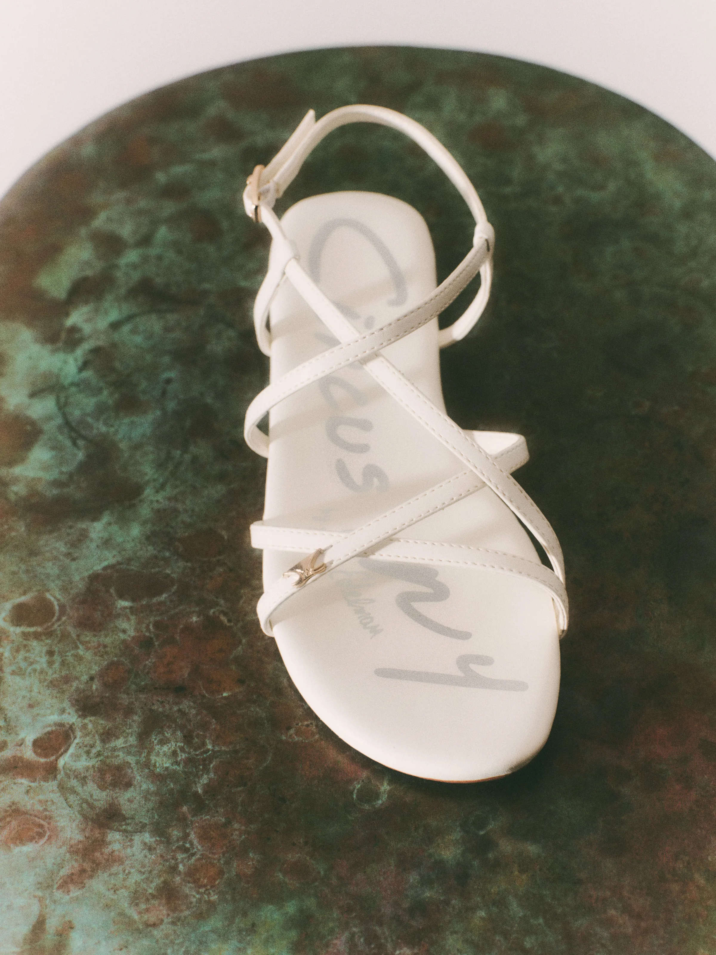 White strappy flat sandal with gold hardware on a green patina metal surface, for Circus NY.