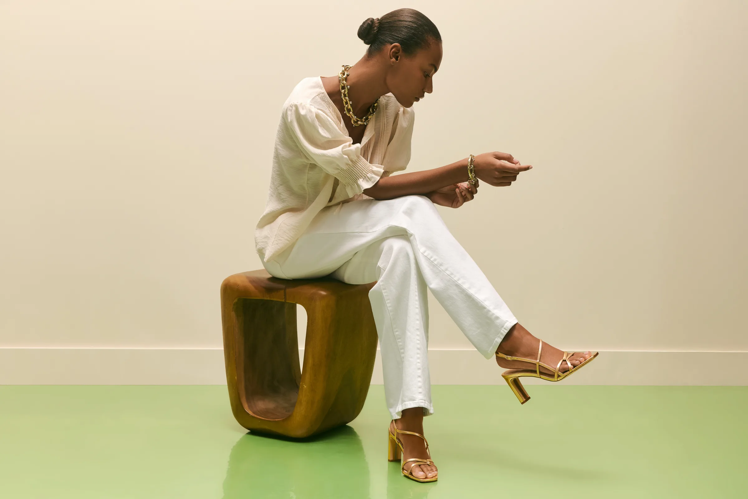 Model in cream blouse and white jeans on a wooden stool, wearing gold strappy heeled sandals, for Sam Edelman.