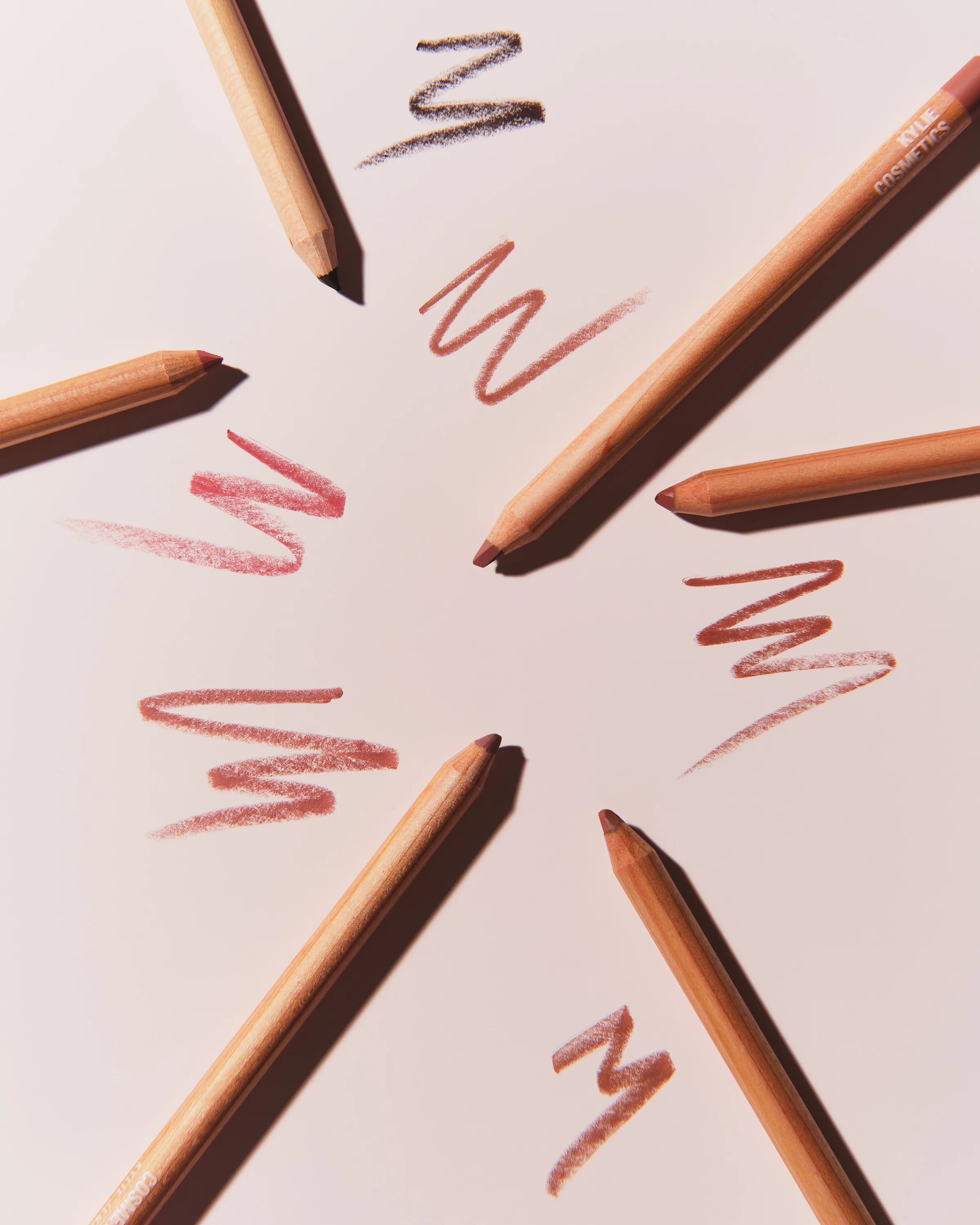 Wooden lip liner pencils arranged in a starburst with pink and brown zigzag swatches on blush paper, for Kylie Cosmetics.