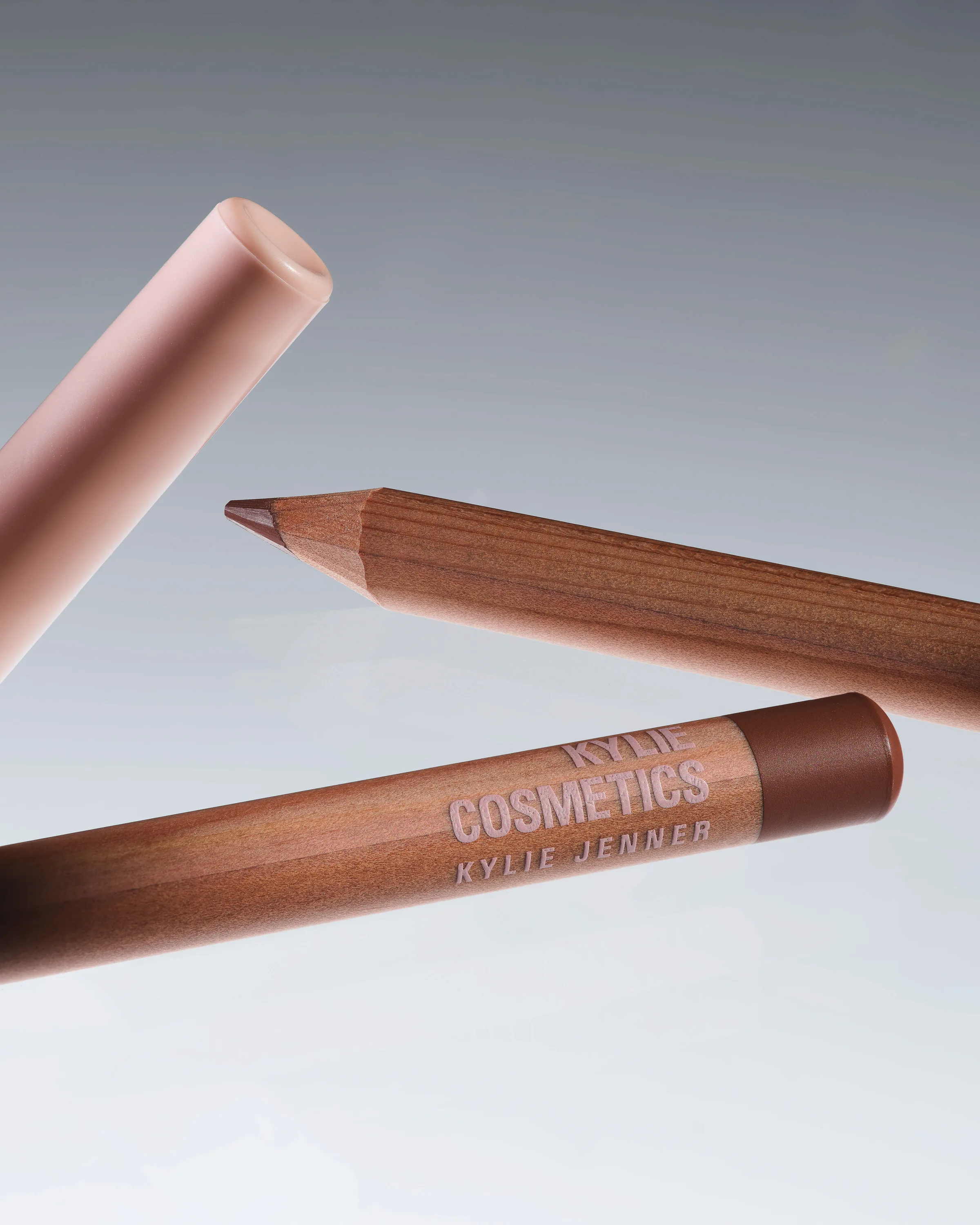 Three wooden lip liner pencils in nude and brown tones floating against a grey gradient backdrop, for Kylie Cosmetics.