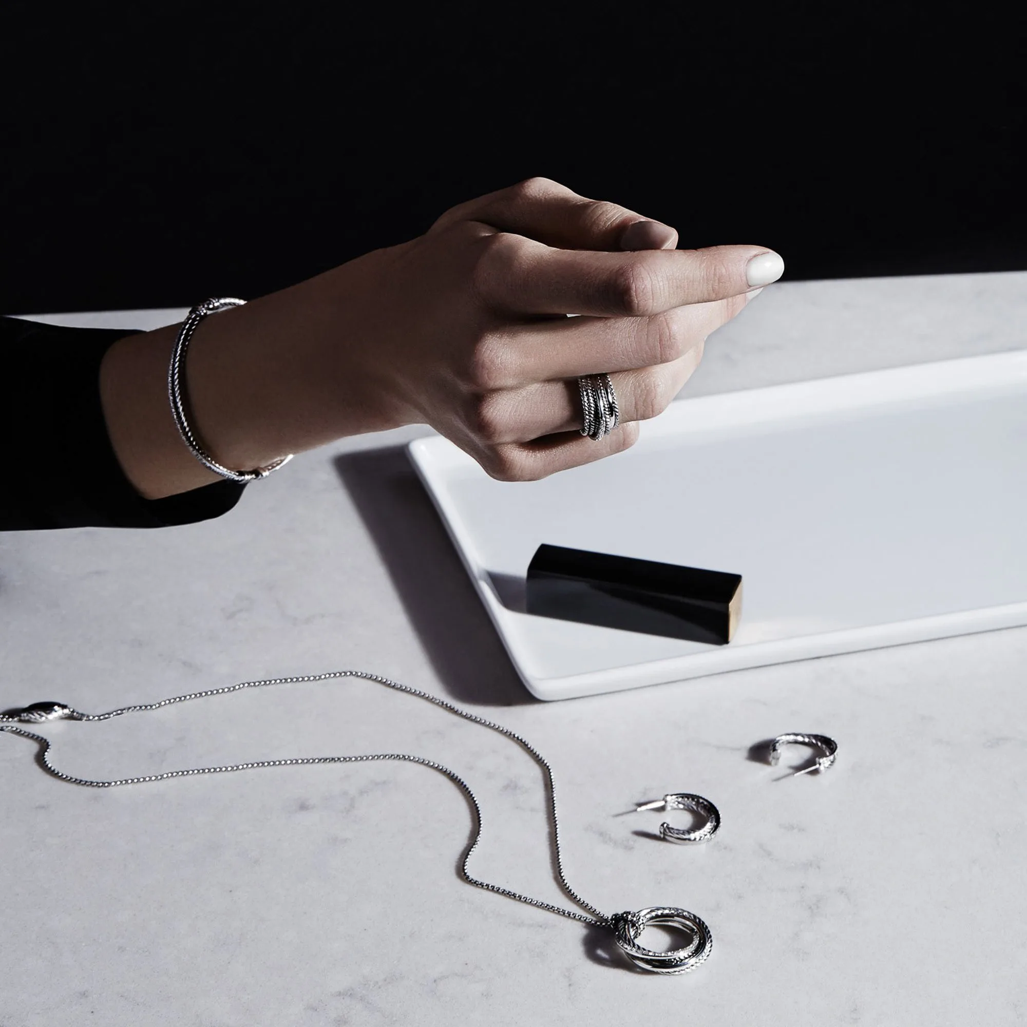 Hand wearing silver cable ring and bracelet above a necklace and hoop earrings on white marble, for David Yurman.