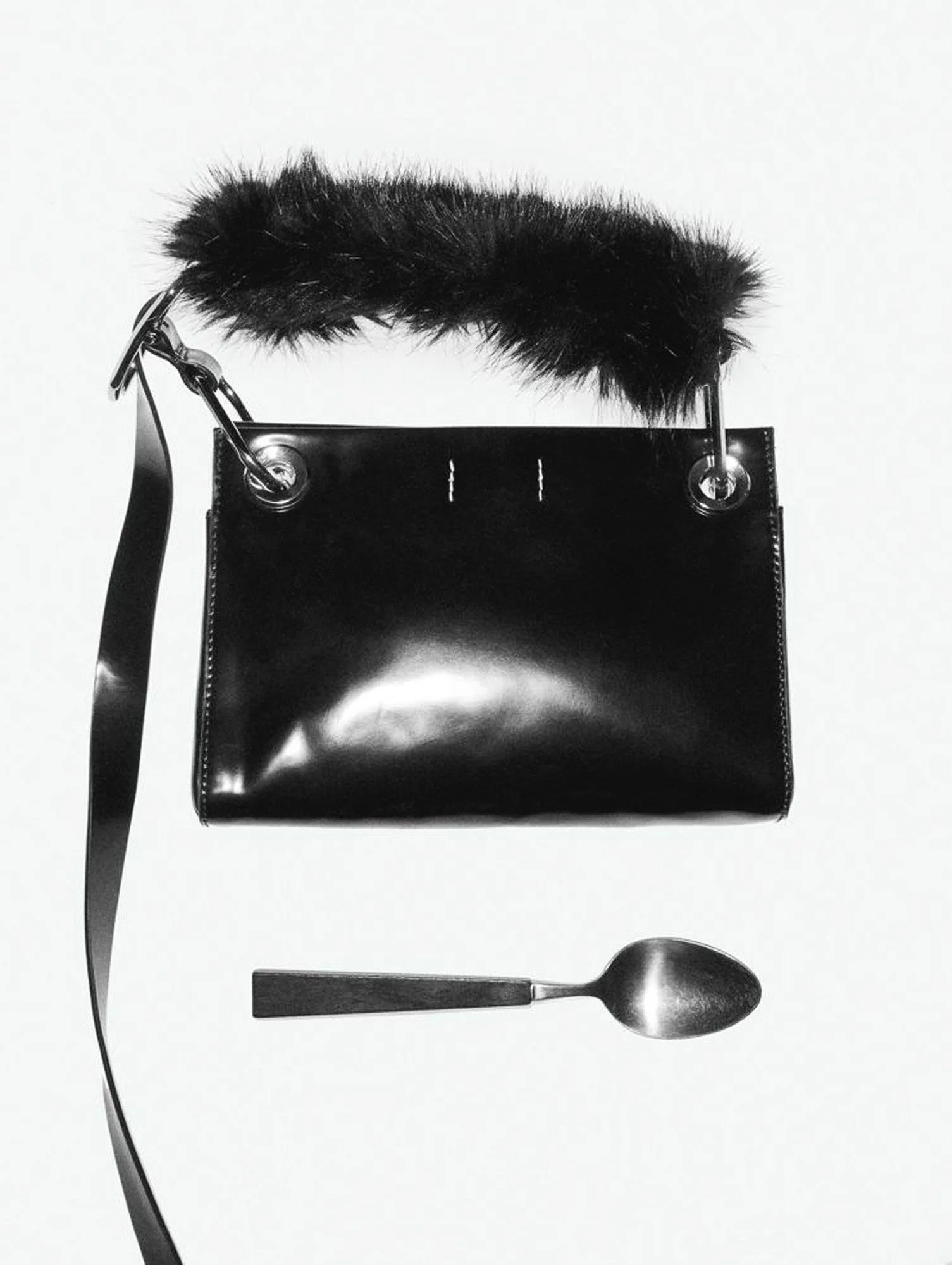 Black patent leather bag with a faux fur handle and long strap beside a steel spoon on white, for Interview Magazine.