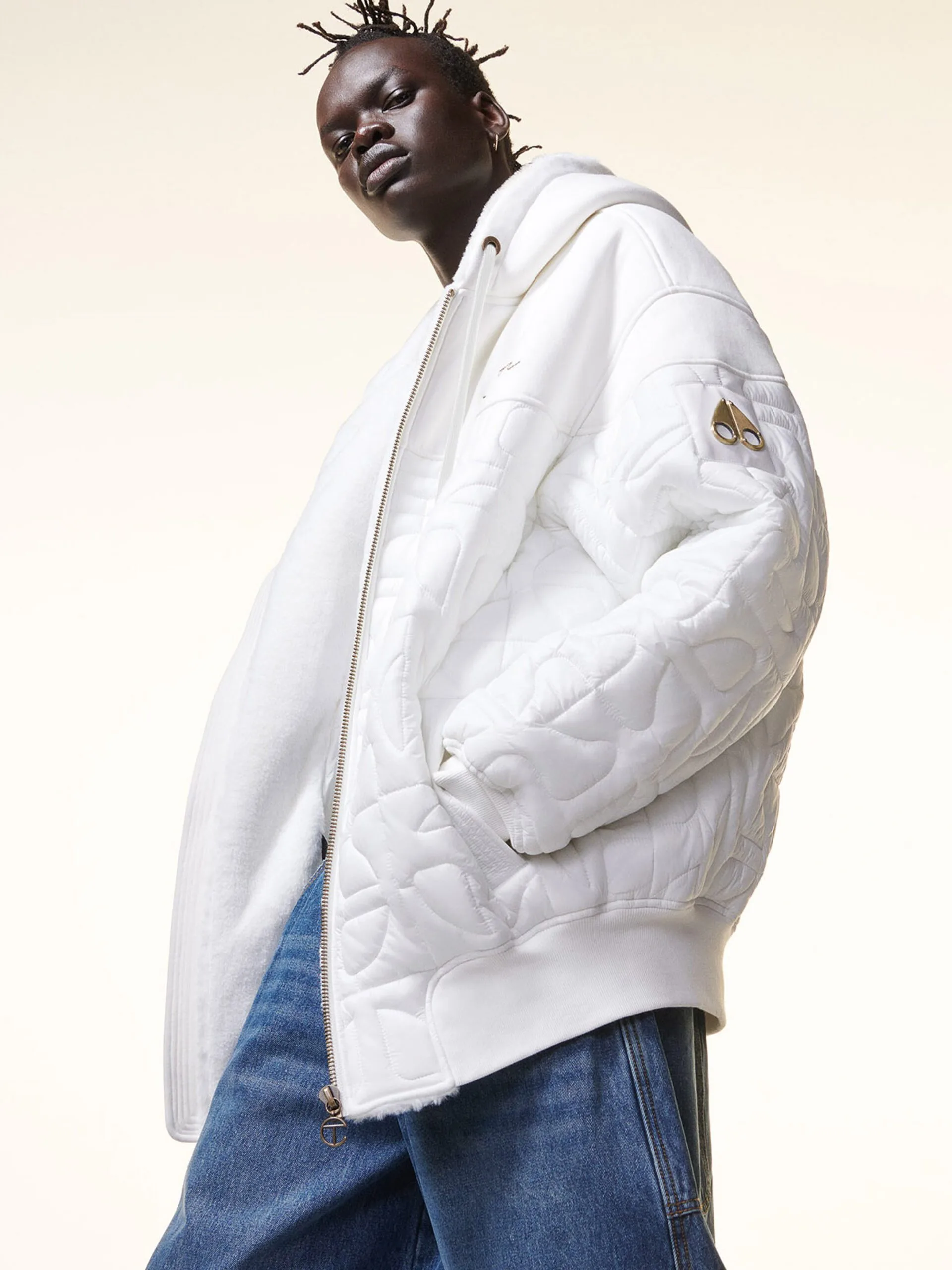 Model in an oversized white quilted hooded jacket with gold logo badge and blue jeans on a cream backdrop, for Moose Knuckles.