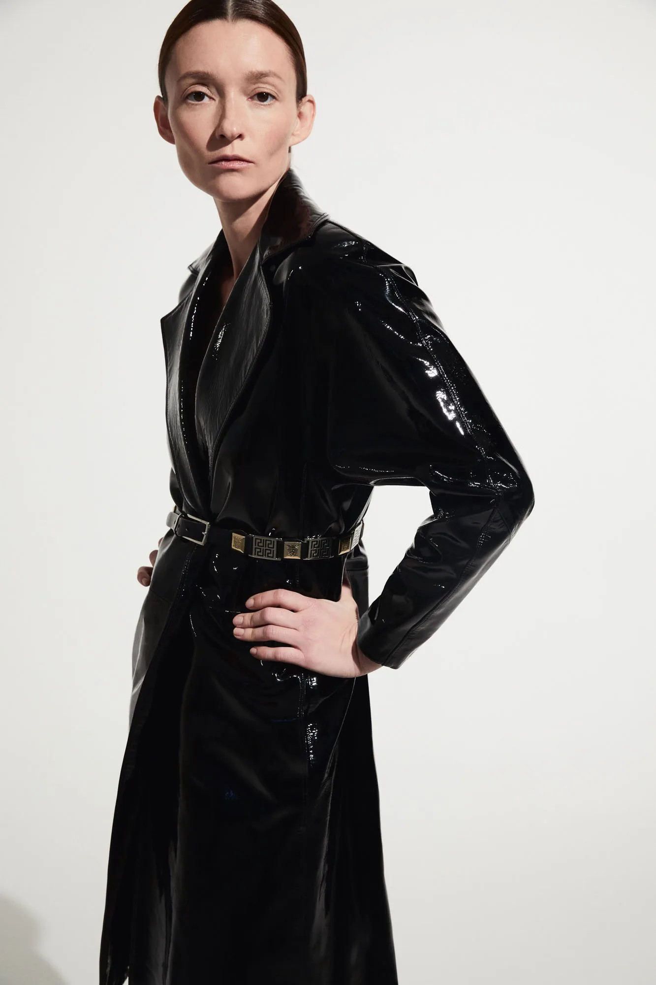Model in a glossy black patent leather trench coat with a metal plaque belt, hand on hip, for The RealReal.