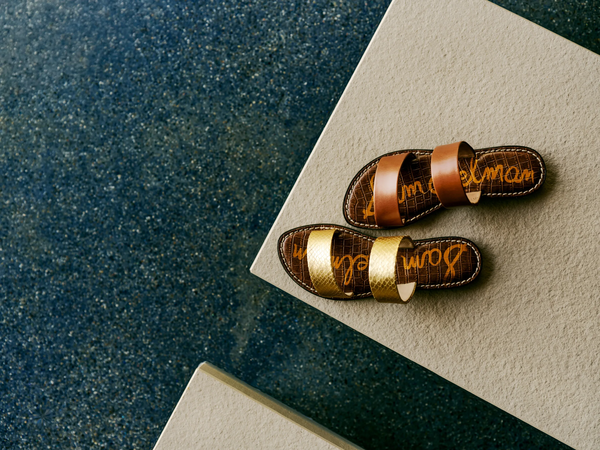 Two double-strap slide sandals, one gold and one tan leather, on a stone ledge above dark terrazzo, for Sam Edelman.