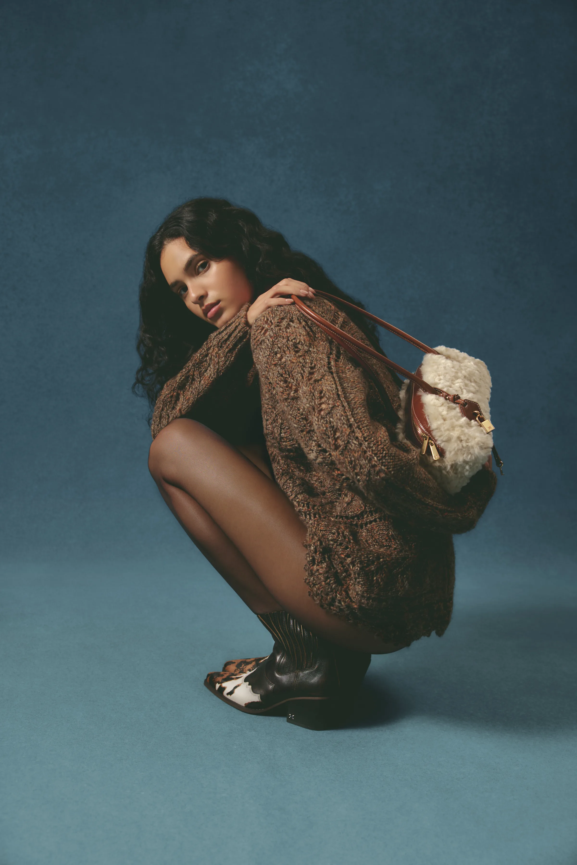 Model crouching in a brown cable knit sweater, cowhide western boots and a shearling shoulder bag on blue, for Sam Edelman.