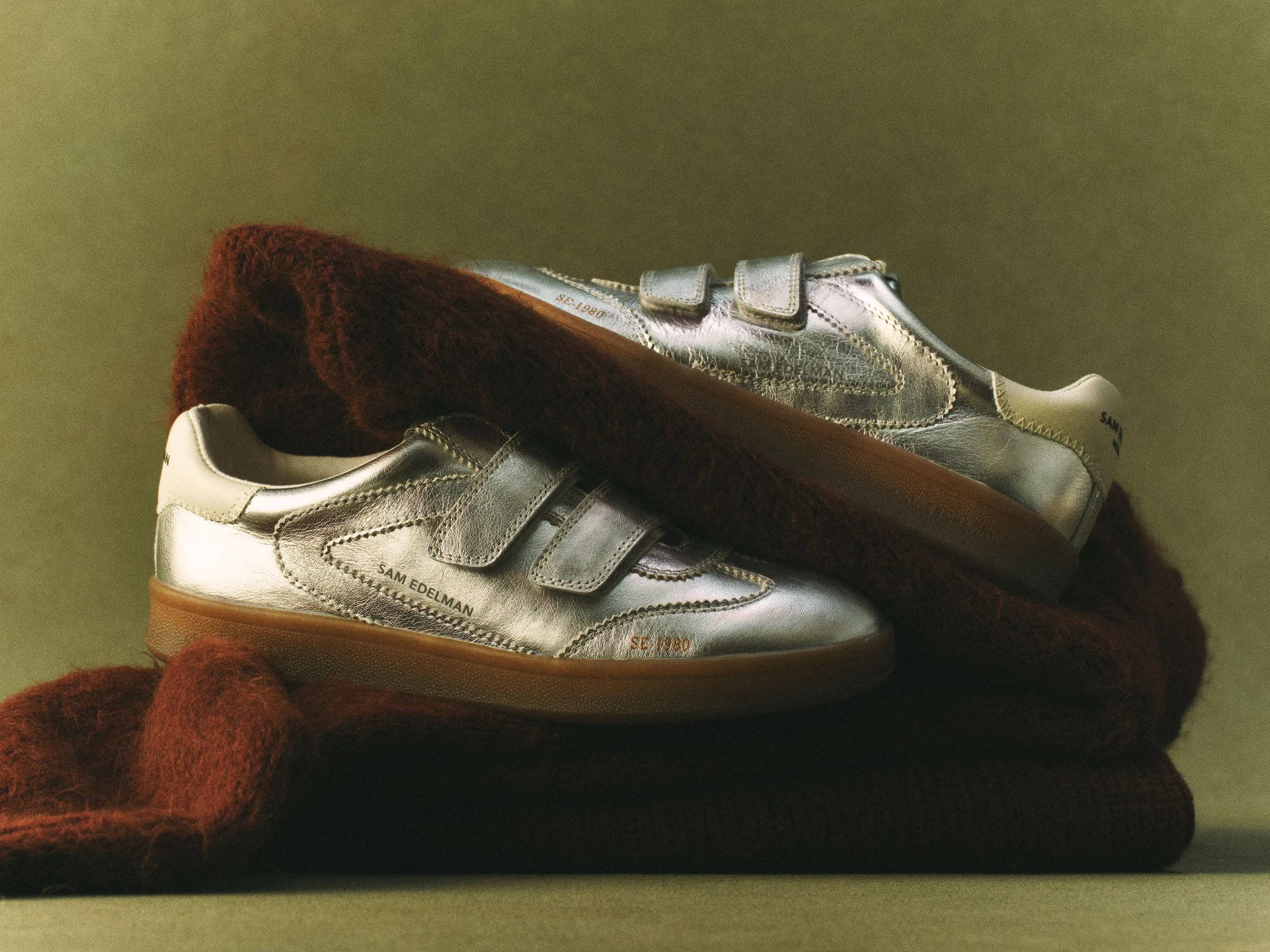 Metallic silver leather sneakers with velcro straps and gum soles resting on a rust mohair knit, olive backdrop, for Sam Edelman.