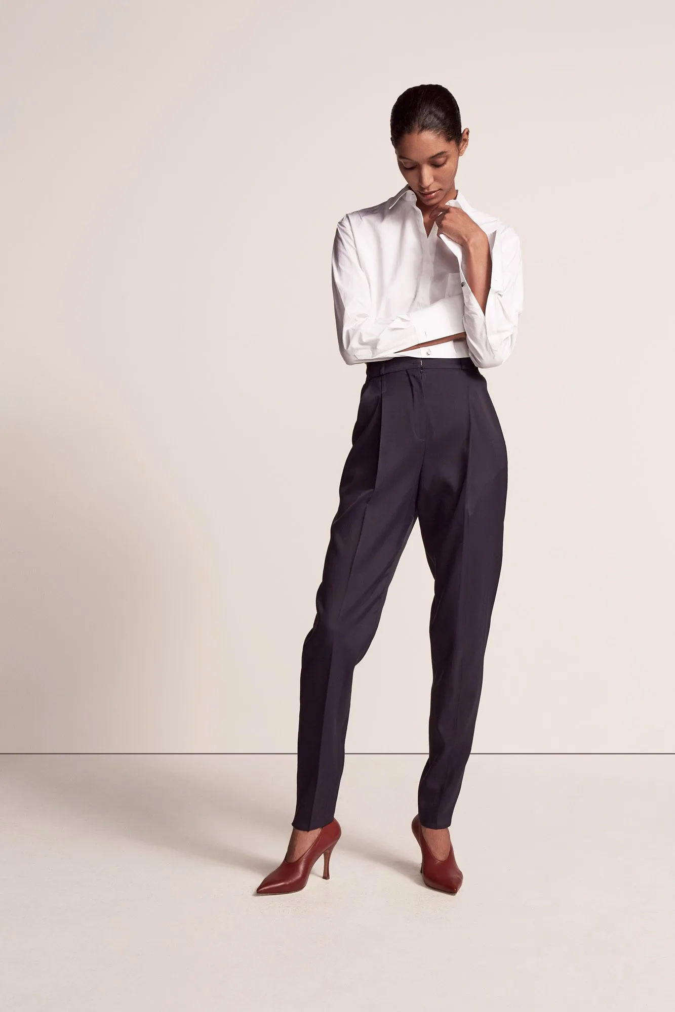 Model in a cropped white shirt, pleated navy trousers and burgundy pumps on a cream backdrop, for Another Tomorrow.