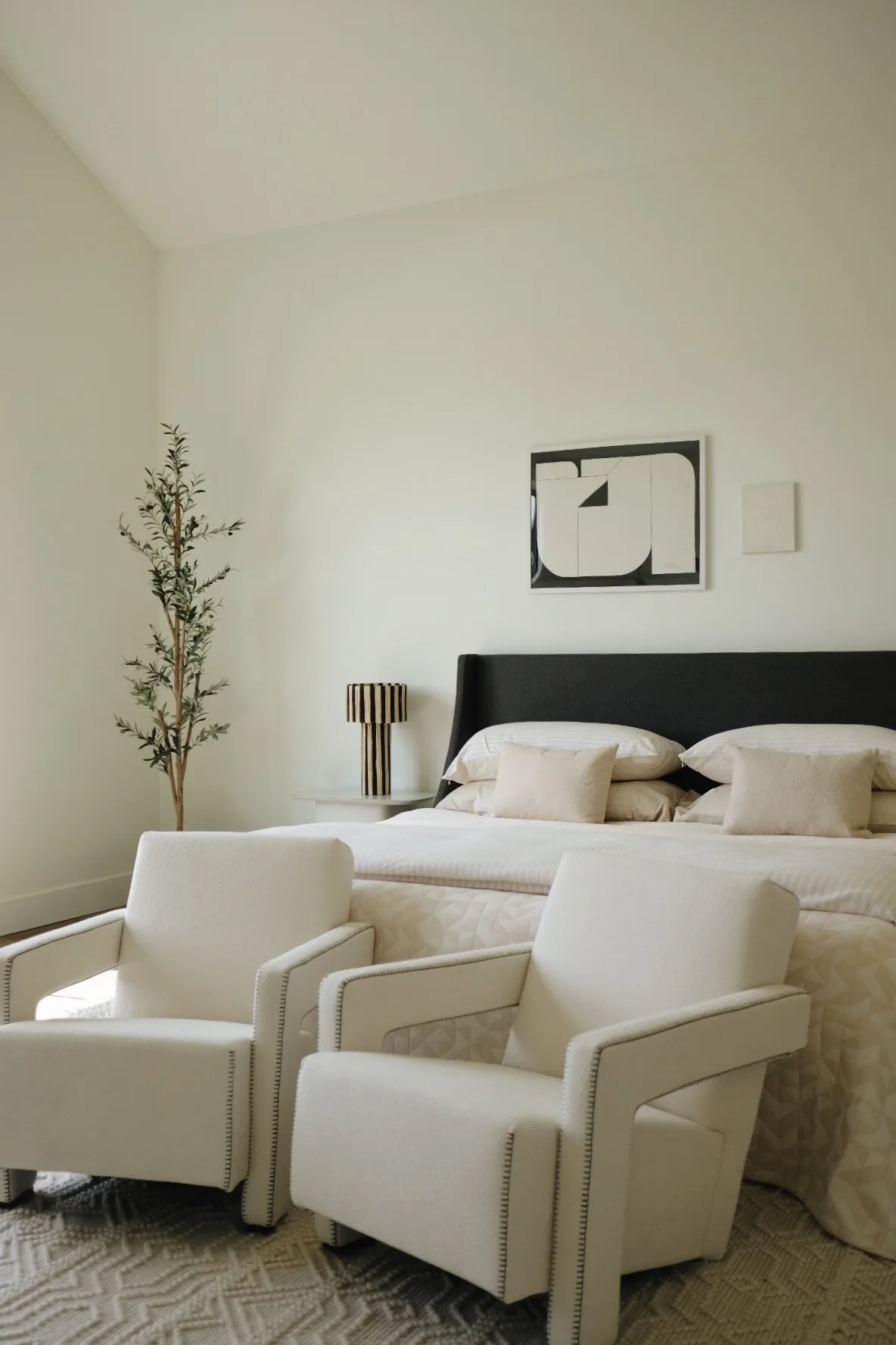 Bedroom with two cream bouclé armchairs, a dark upholstered headboard, quilted bedding and an olive tree, for Jennifer Hagler.