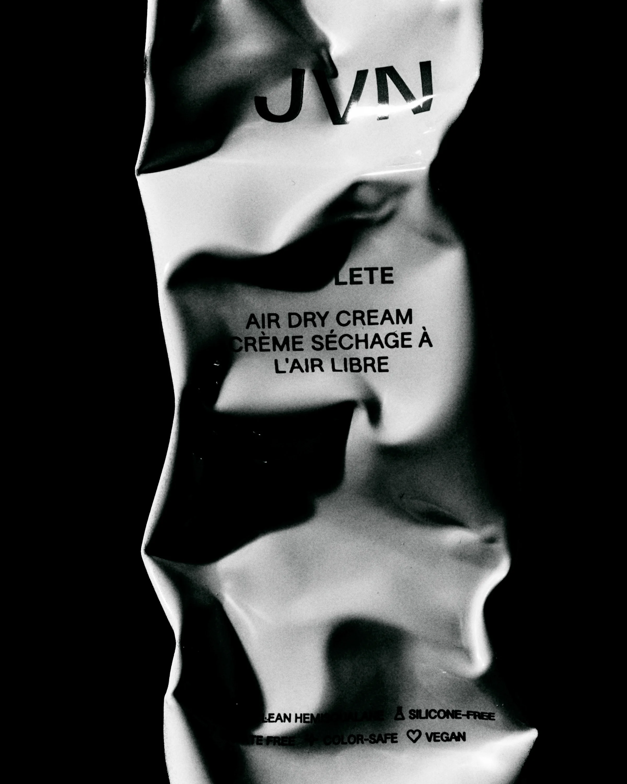 Black and white close-up of a crumpled metallic Complete Air Dry Cream tube against a black background, for JVN.