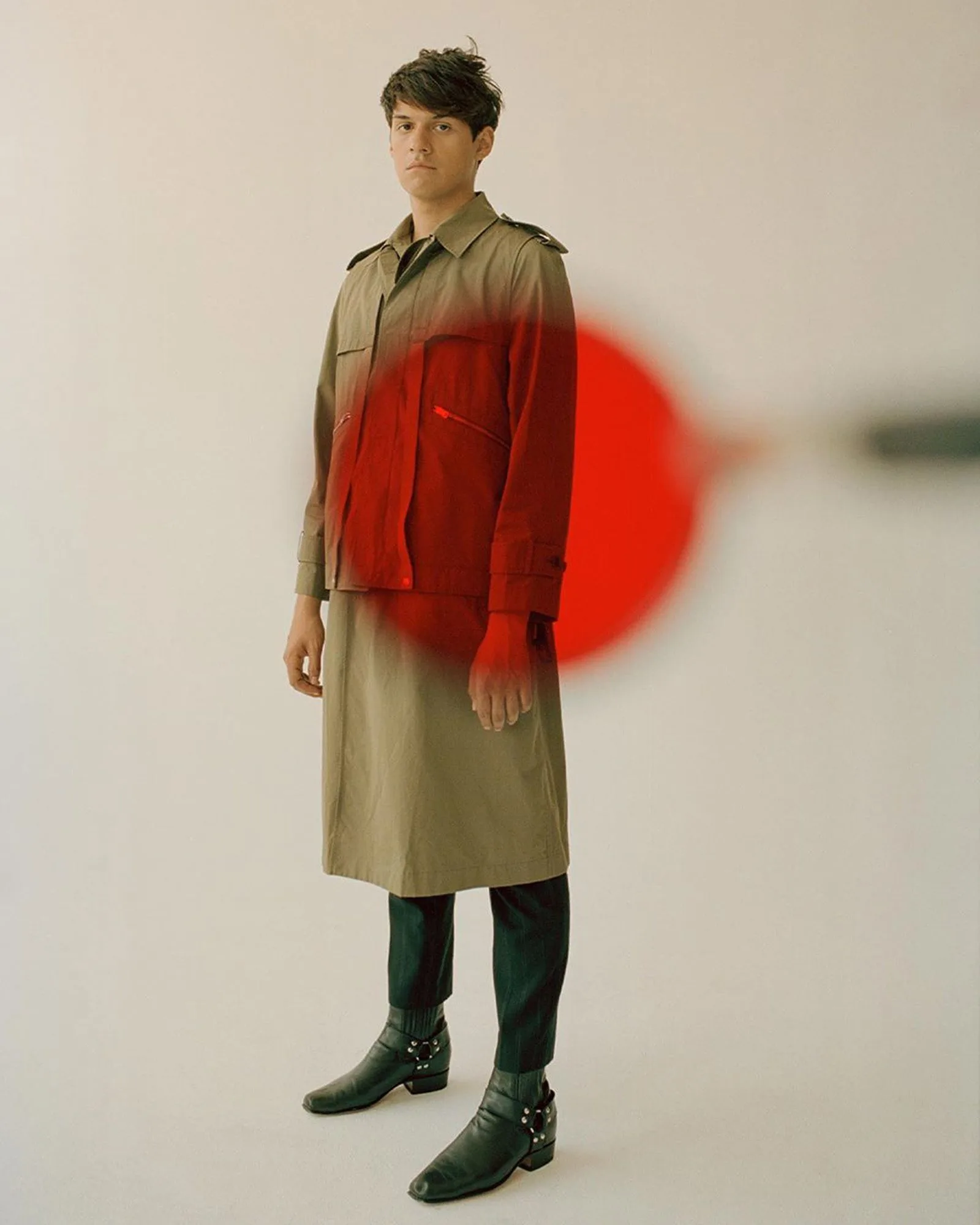 Man in a khaki trench coat over a skirt-length layer and black harness boots, a blurred red disc in front, for The Last Magazine.