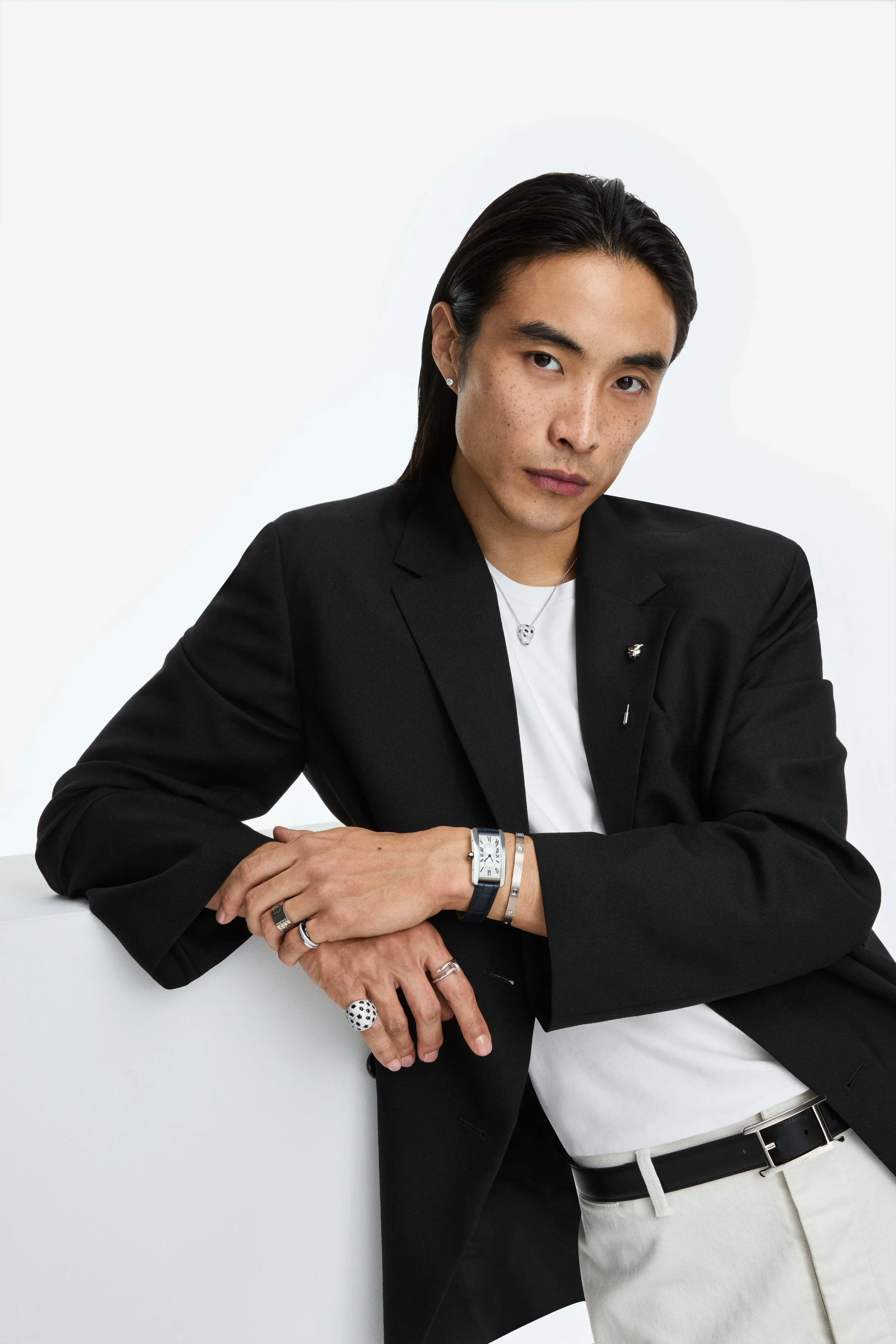 Model in a black blazer and white tee leaning on a white block, wearing stacked rings, bracelets and a watch, for Cartier.