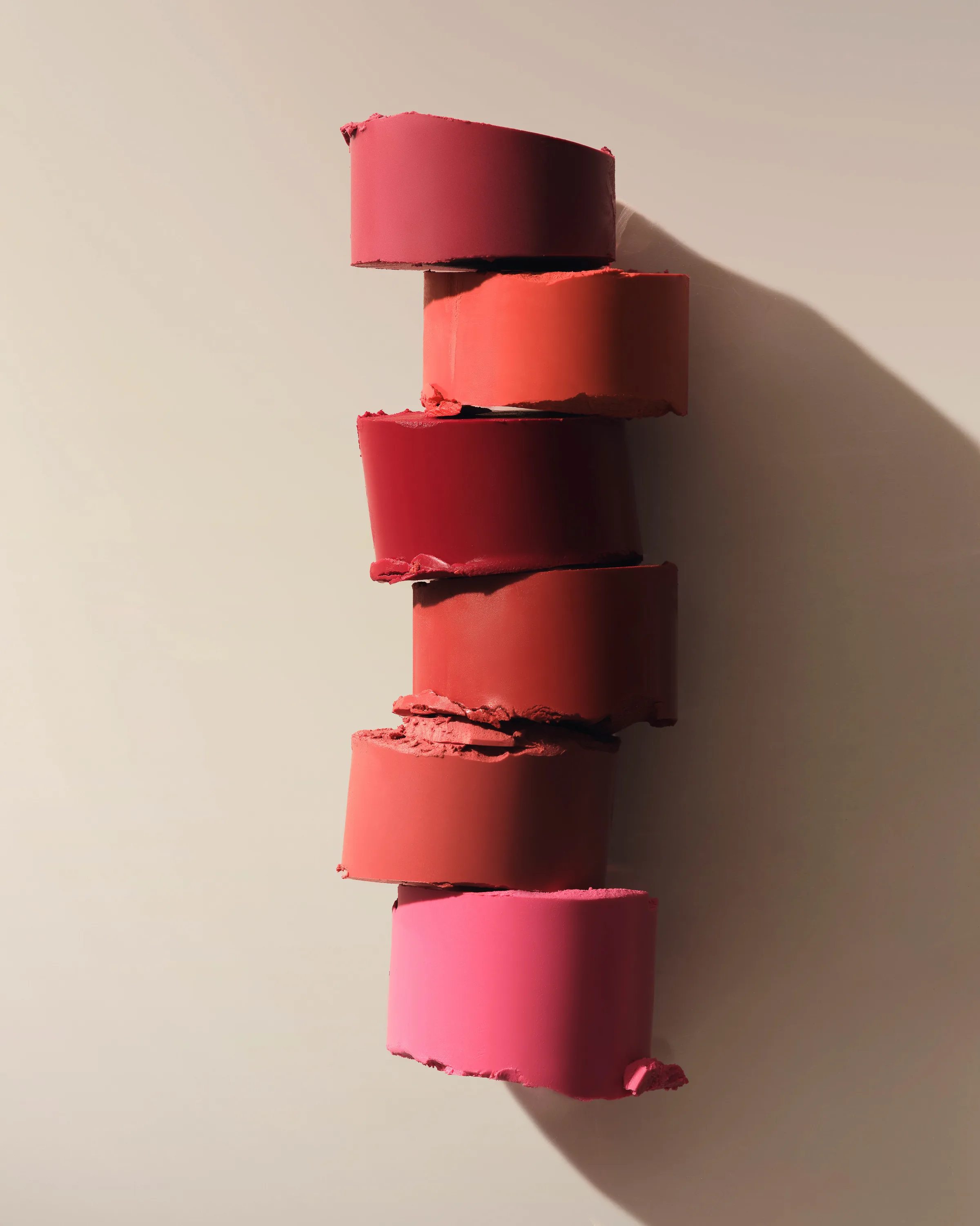 Still life of six sliced lipstick bullets in red, coral and pink stacked against a beige wall, for Kylie Cosmetics.