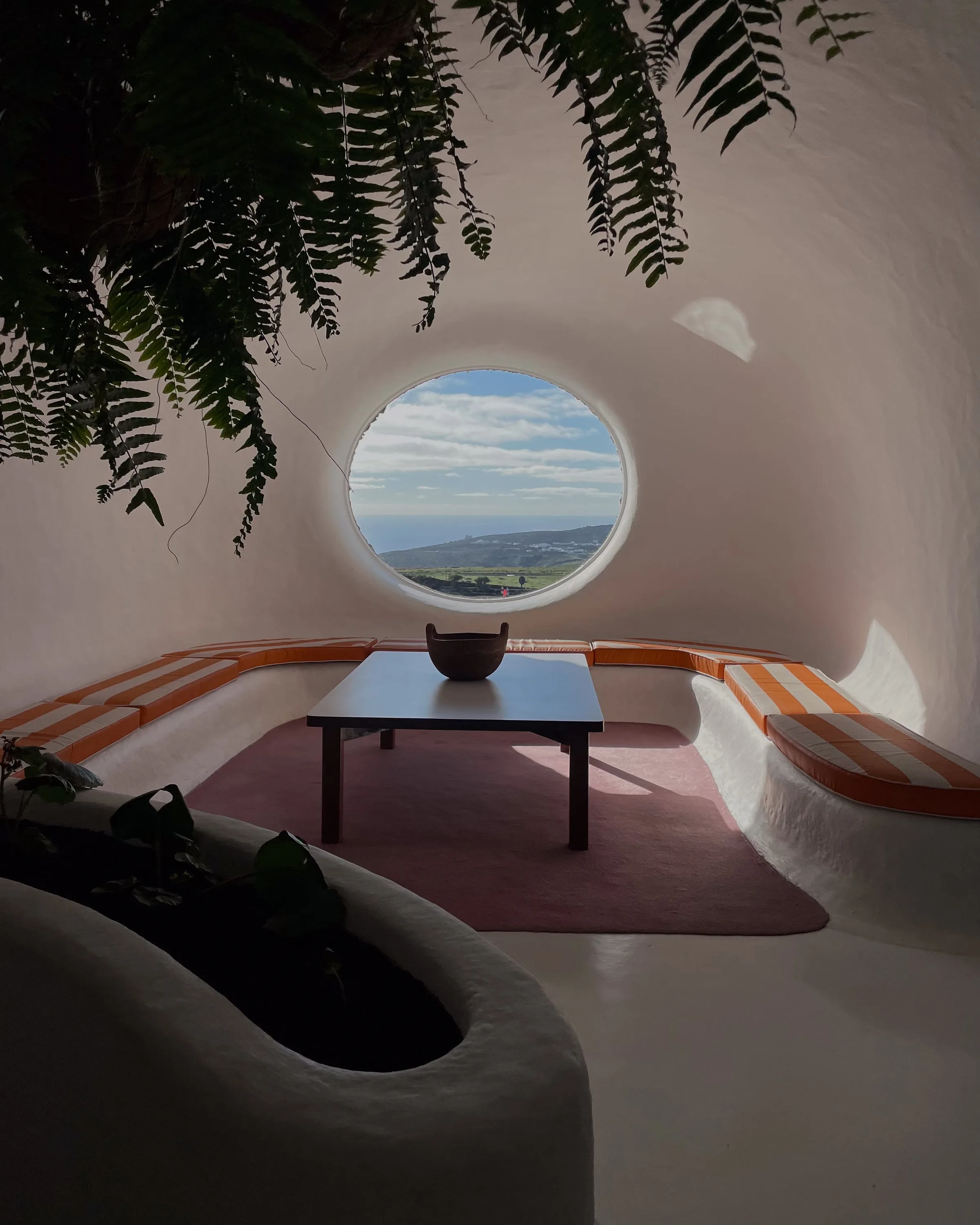 Whitewashed cave lounge with orange striped bench cushions, low table and round window overlooking hills, ferns above.