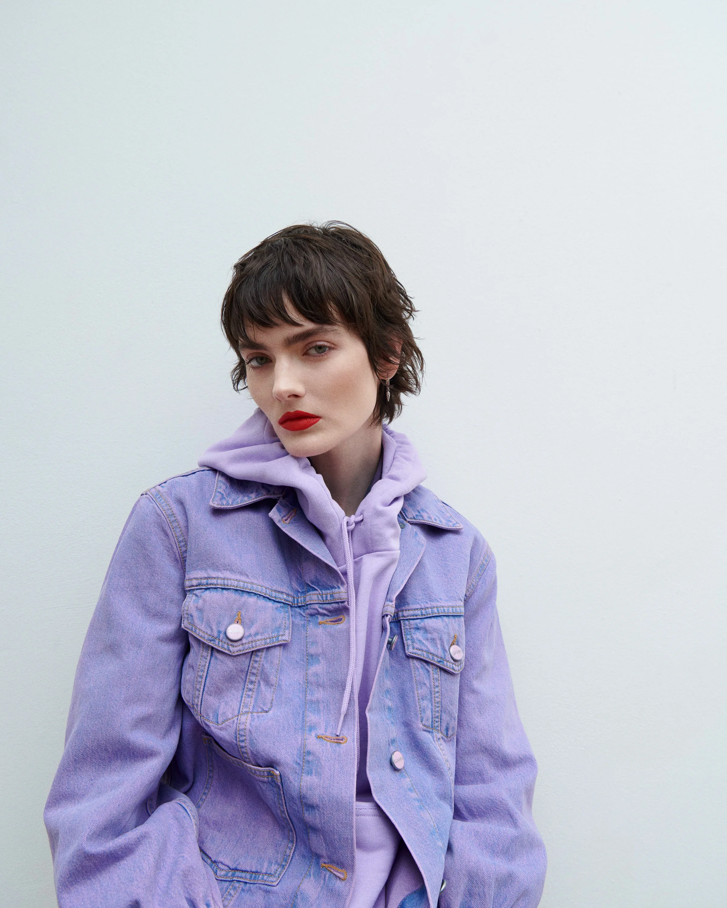 Model with short dark hair and red lips in a lavender denim jacket and hoodie against a white wall.