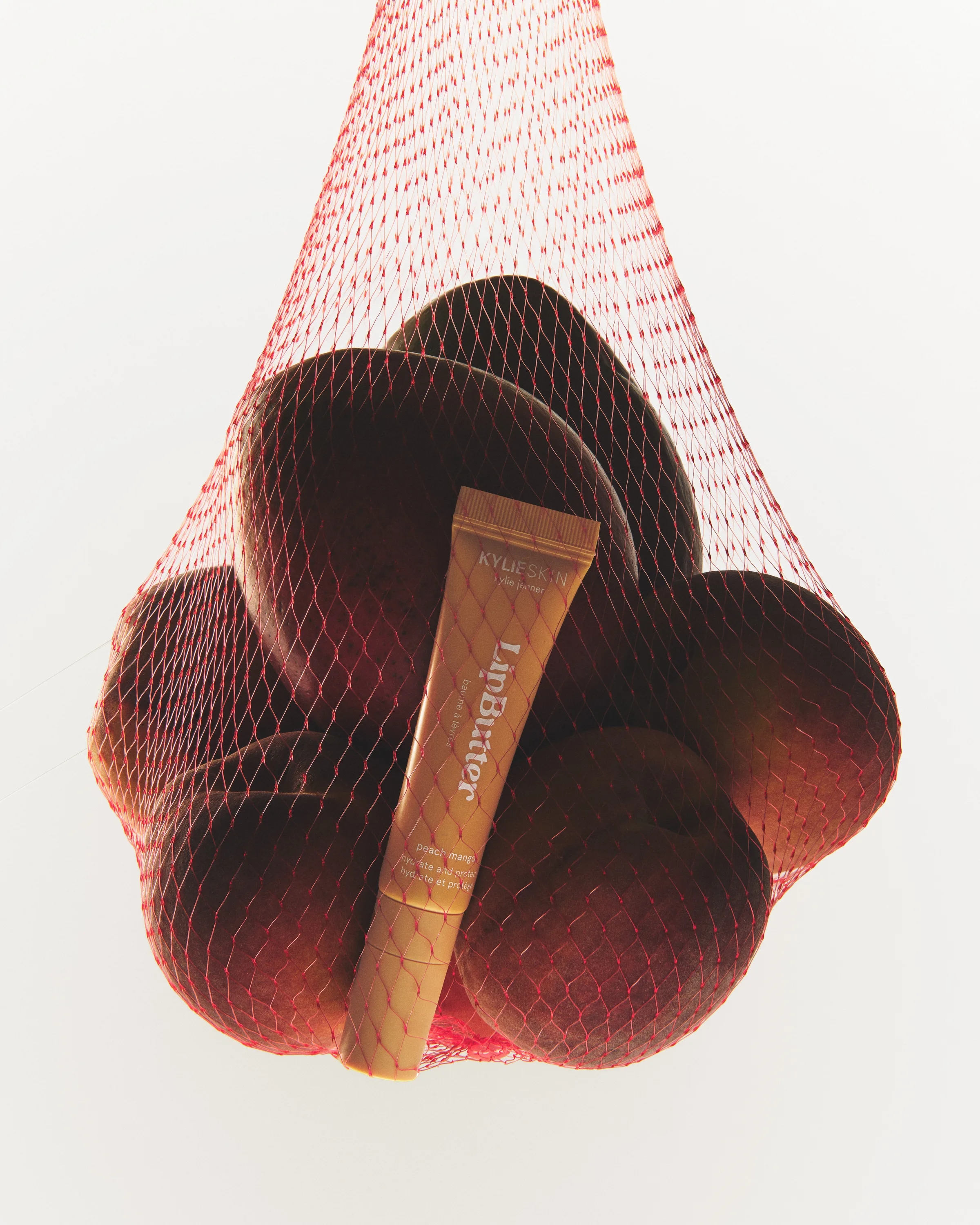Peach mango Lip Butter tube tucked among peaches in a red mesh produce bag against a white backdrop, for Kylie Cosmetics.