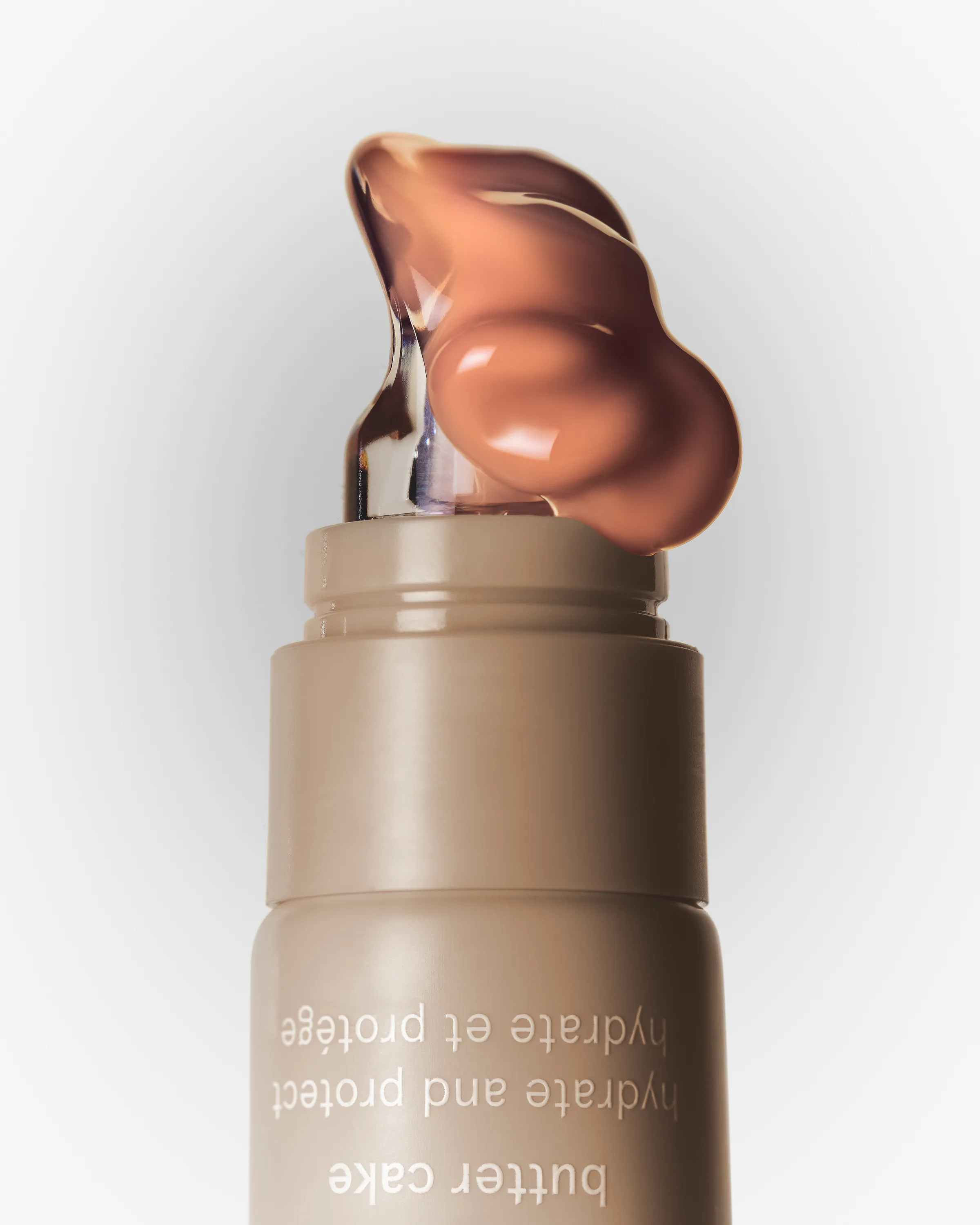 Still life of a tan lip butter tube in Butter Cake with a glossy dollop of balm on its tip, for Kylie Cosmetics.