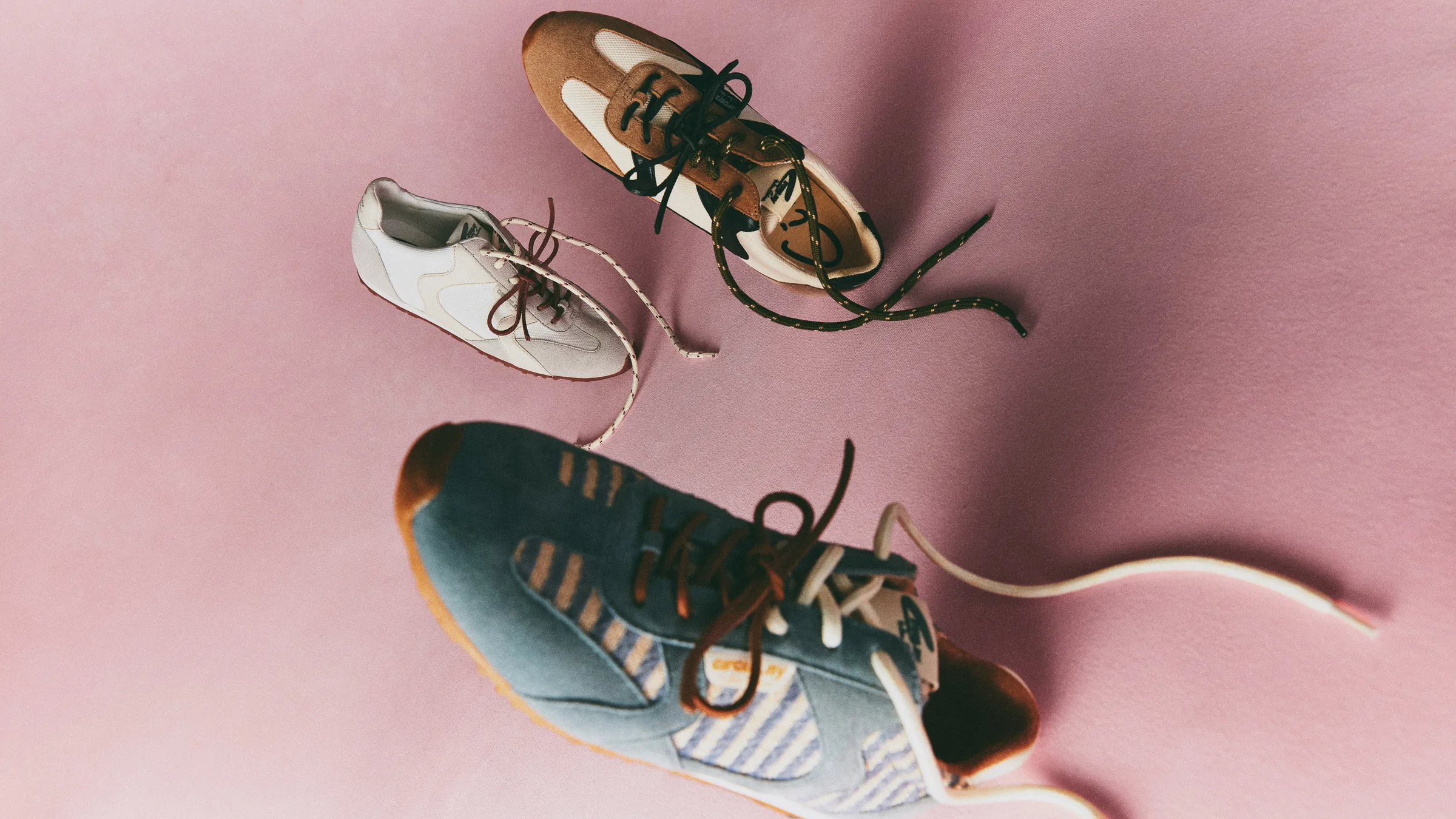 Three Nash retro sneakers in white, tan suede and blue denim scattered with loose laces on a pink backdrop, for Sam Edelman.