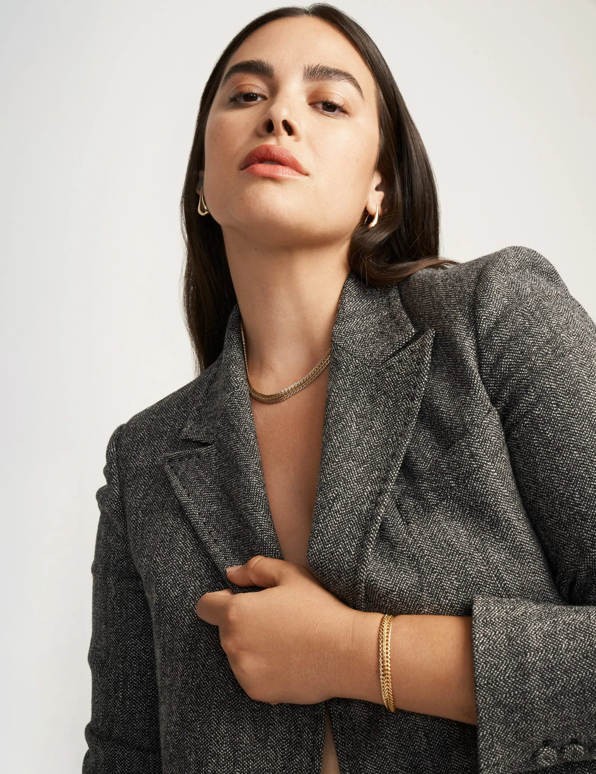 Model in a grey herringbone tweed blazer with gold chain necklace, hoops and bracelet on a white backdrop, for Anne Klein.