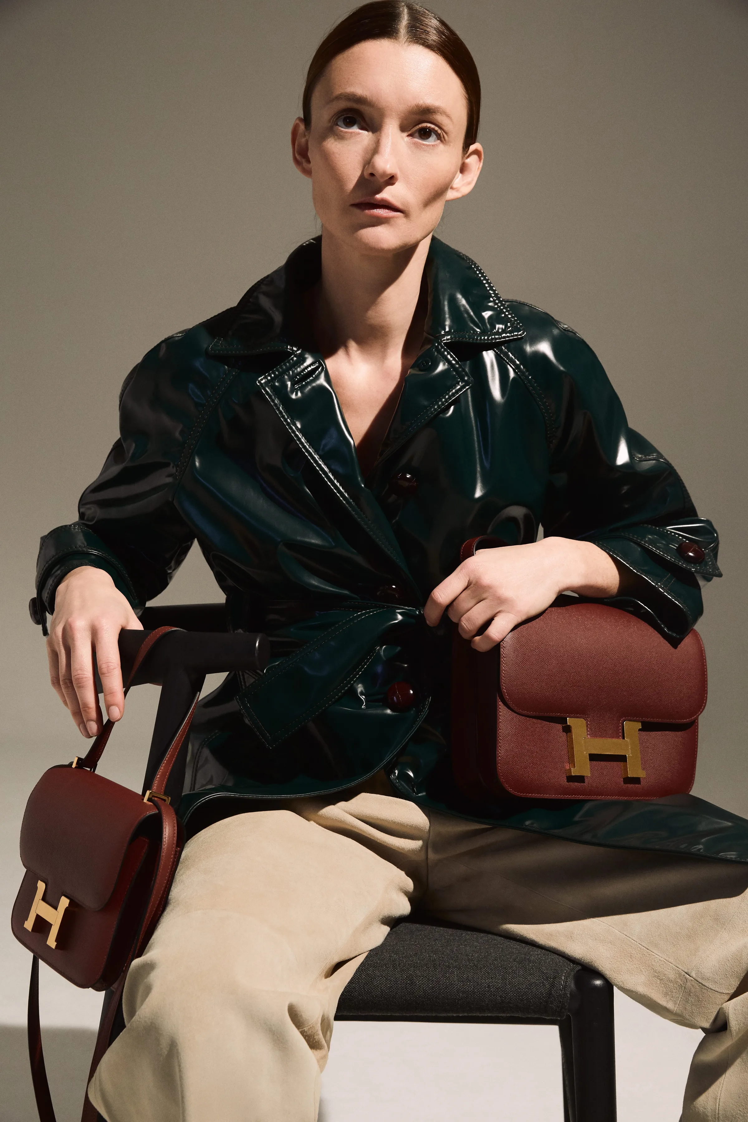 Model in a glossy green patent trench and beige trousers seated holding two burgundy Hermès Constance bags, for The RealReal.