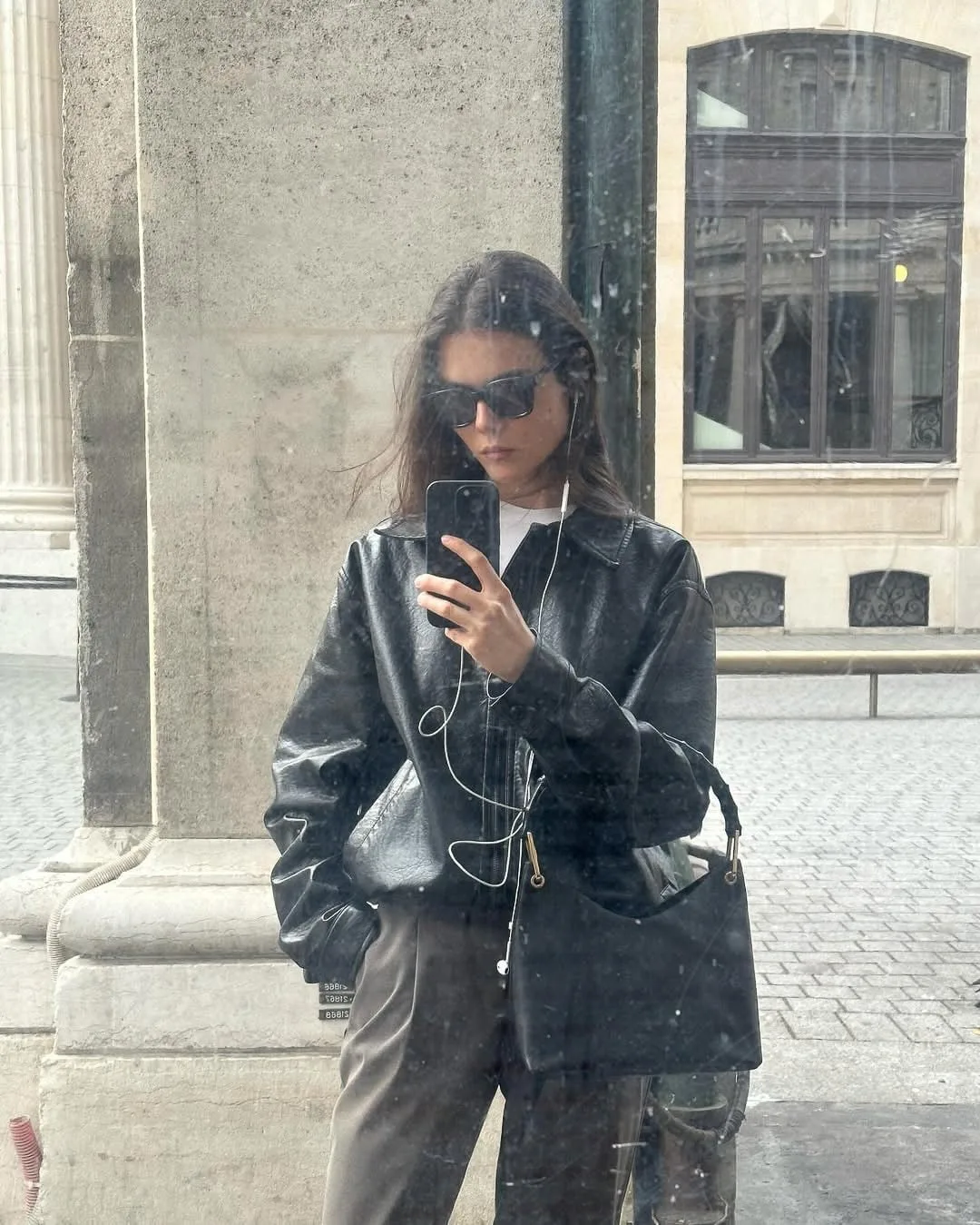 Woman in sunglasses and black leather jacket takes a phone mirror selfie on a city street, carrying a black shoulder bag.