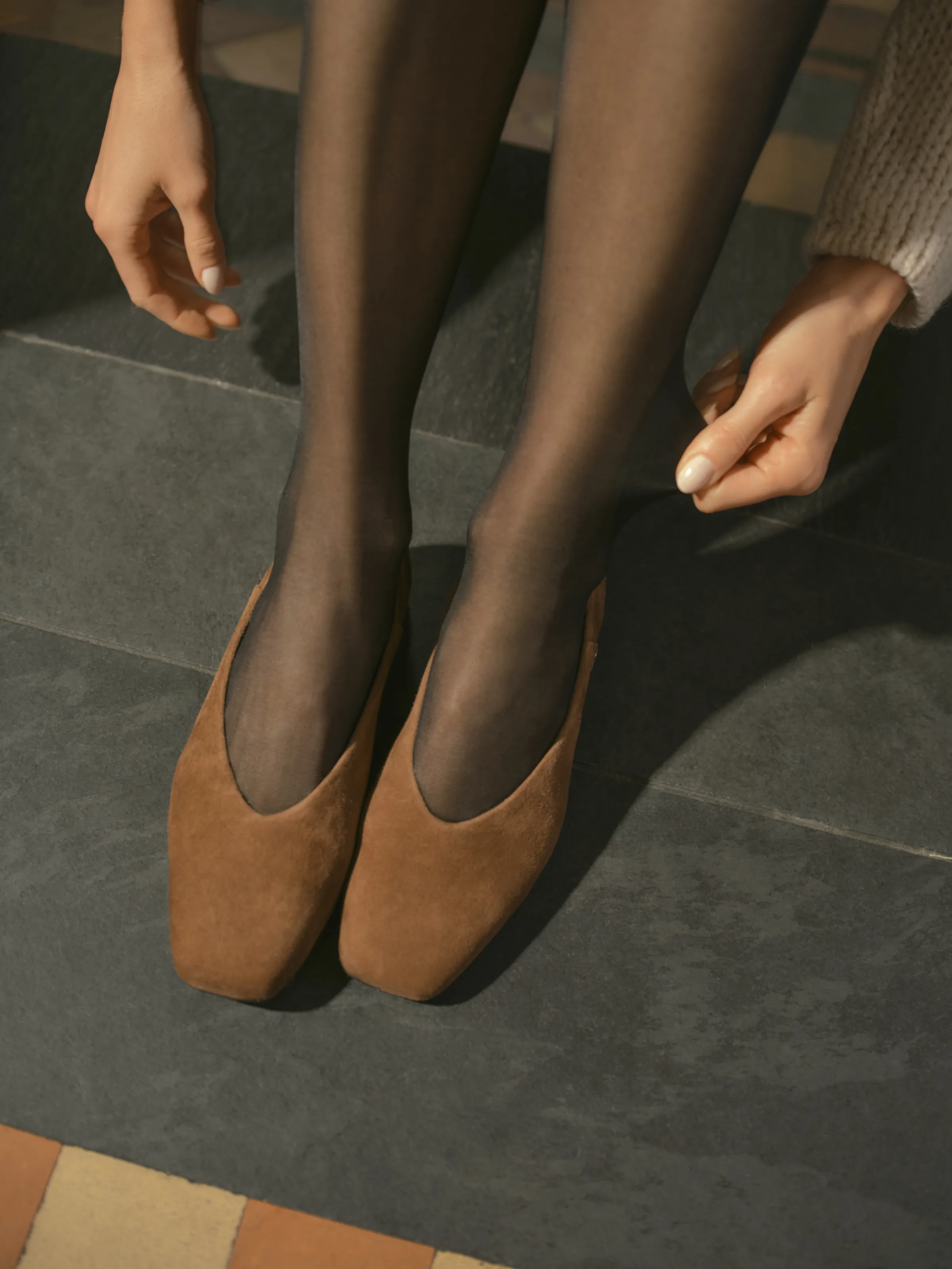 Tan suede square-toe flats worn with sheer black tights on a slate tile floor, hands reaching down, for Sam Edelman.