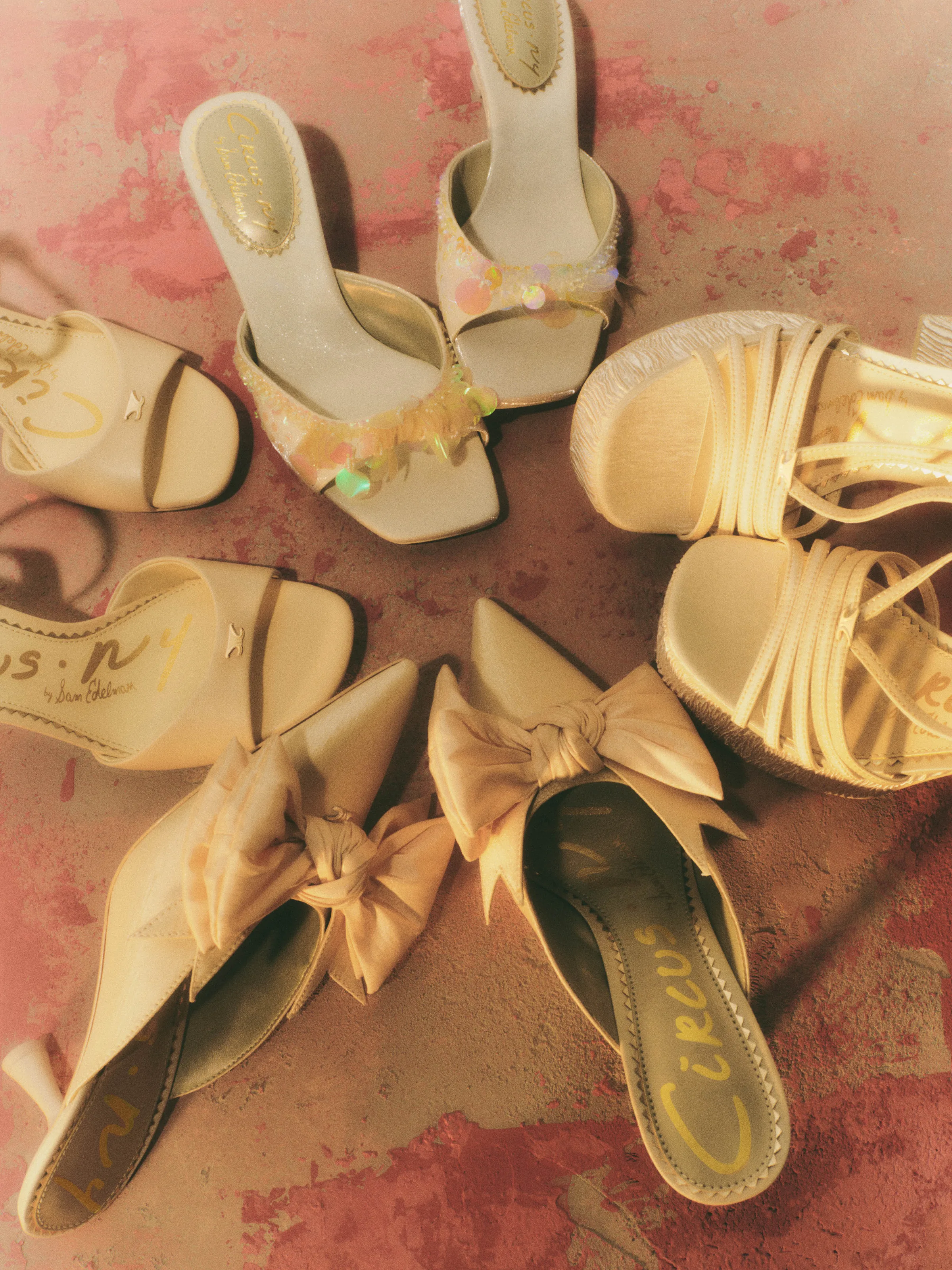 Cream and pearl sandals, bow mules and sequin heels arranged in a circle on a pink weathered floor, for Circus NY.