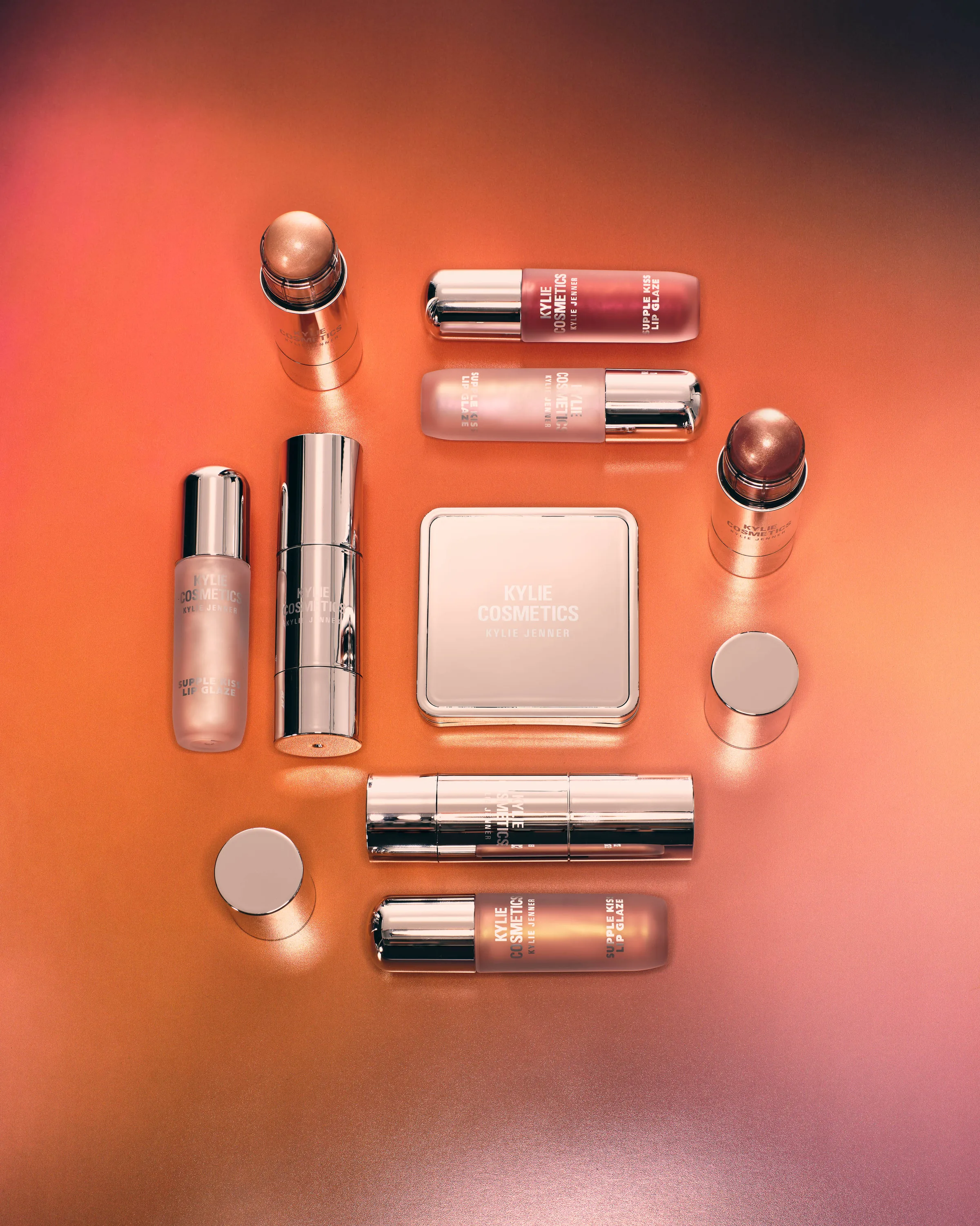 Flat lay of chrome and pink lip glazes, blush sticks and a compact on an orange gradient backdrop, for Kylie Cosmetics.