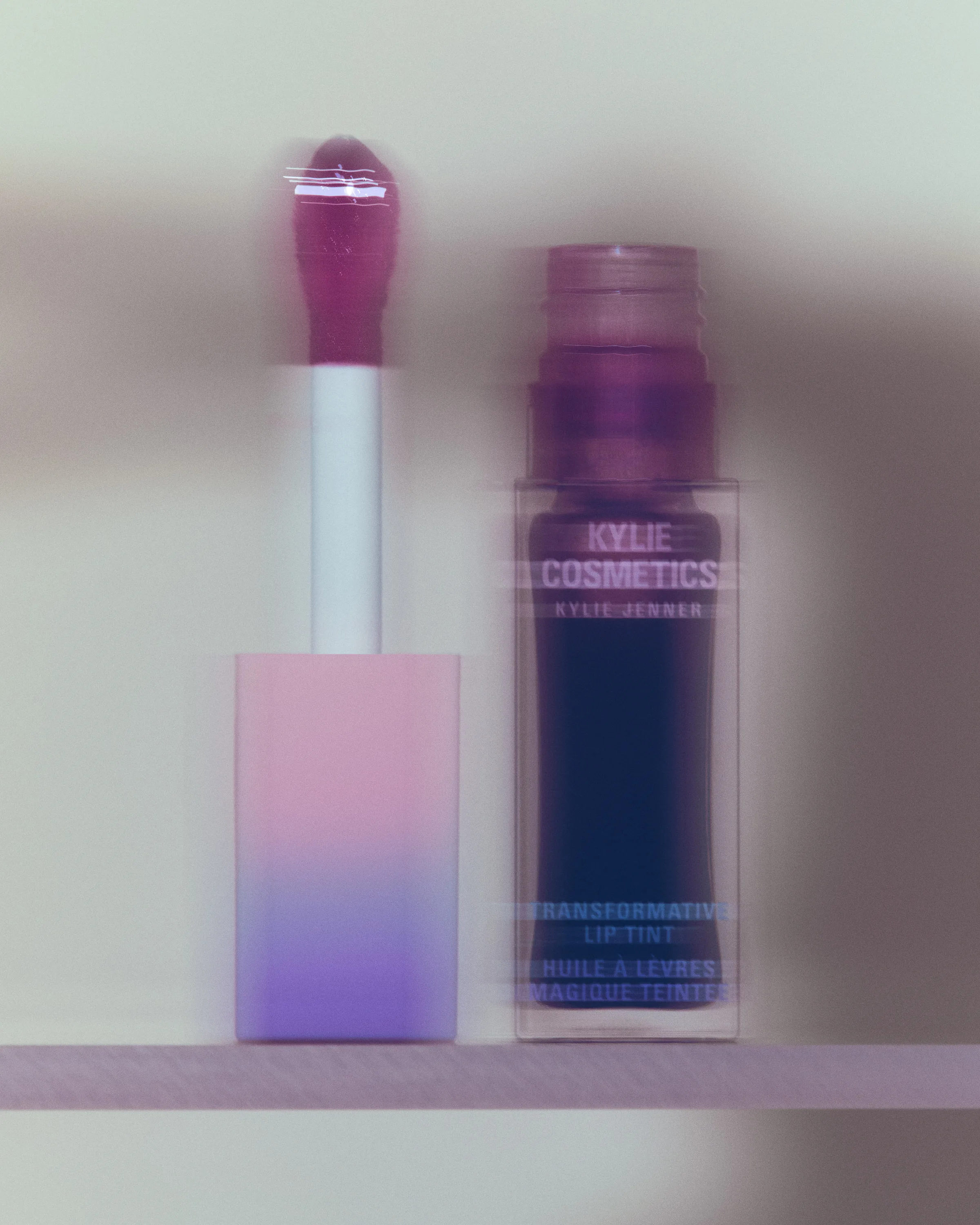 Plum transformative lip tint bottle beside its pink-to-lilac applicator on a shelf, with motion blur, for Kylie Cosmetics.
